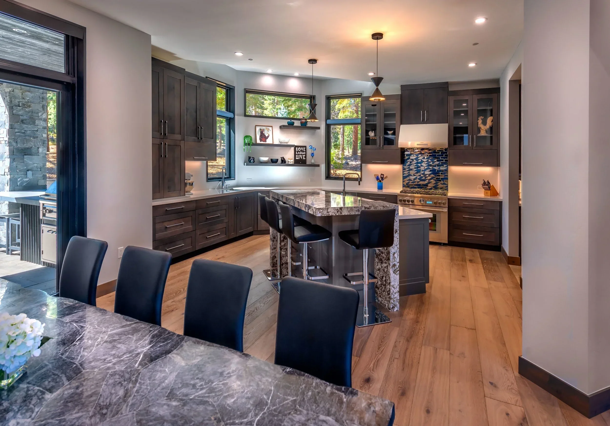 Modern kitchen with dark gray cabinets, a marble-topped island with seating, large windows, and hardwood floors.