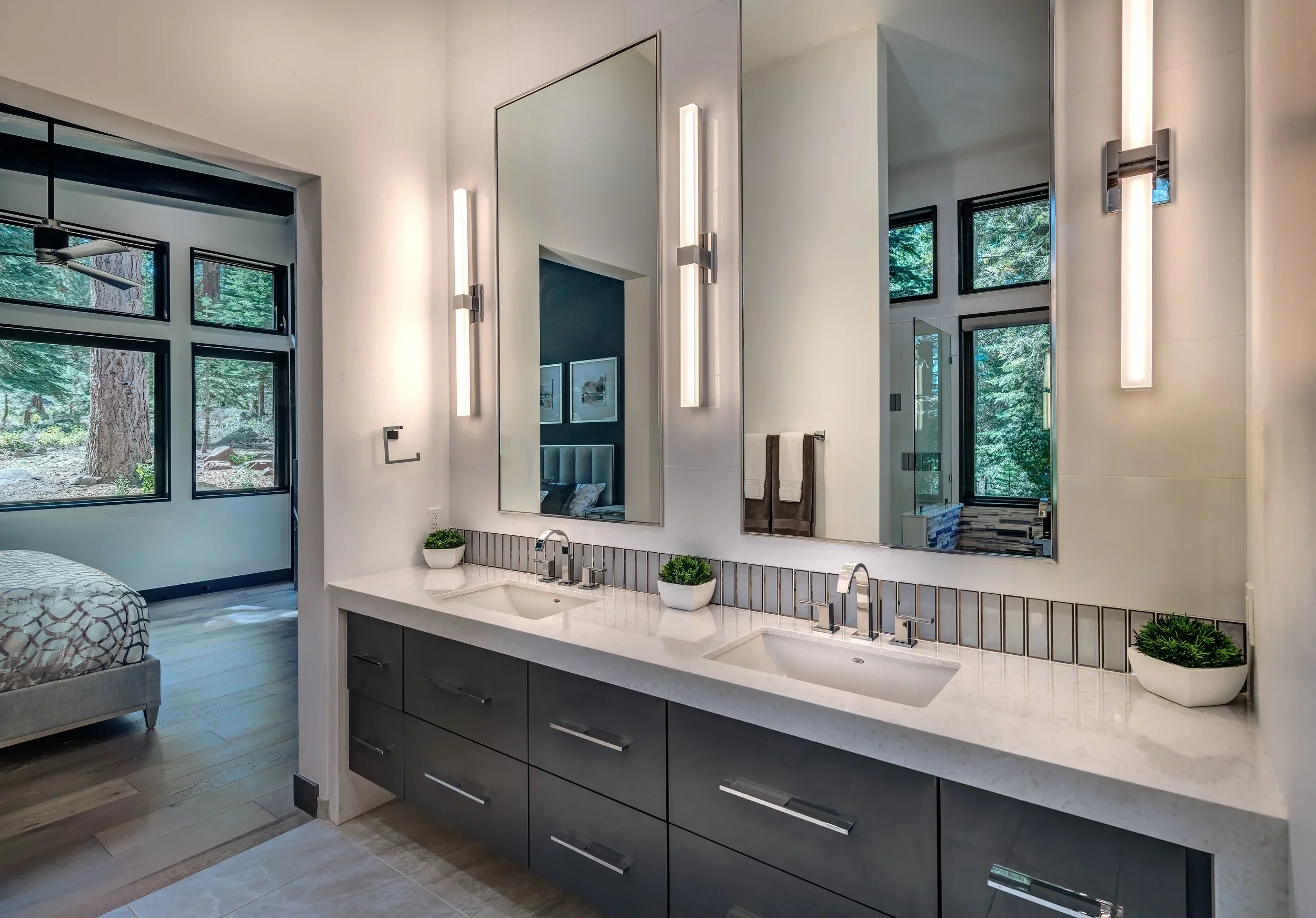 Modern bathroom with a double vanity, large mirrors, vertical lighting fixtures, and two small potted plants on a white countertop. A bedroom with a bed and large windows overlooking a wooded area is visible through an open doorway.