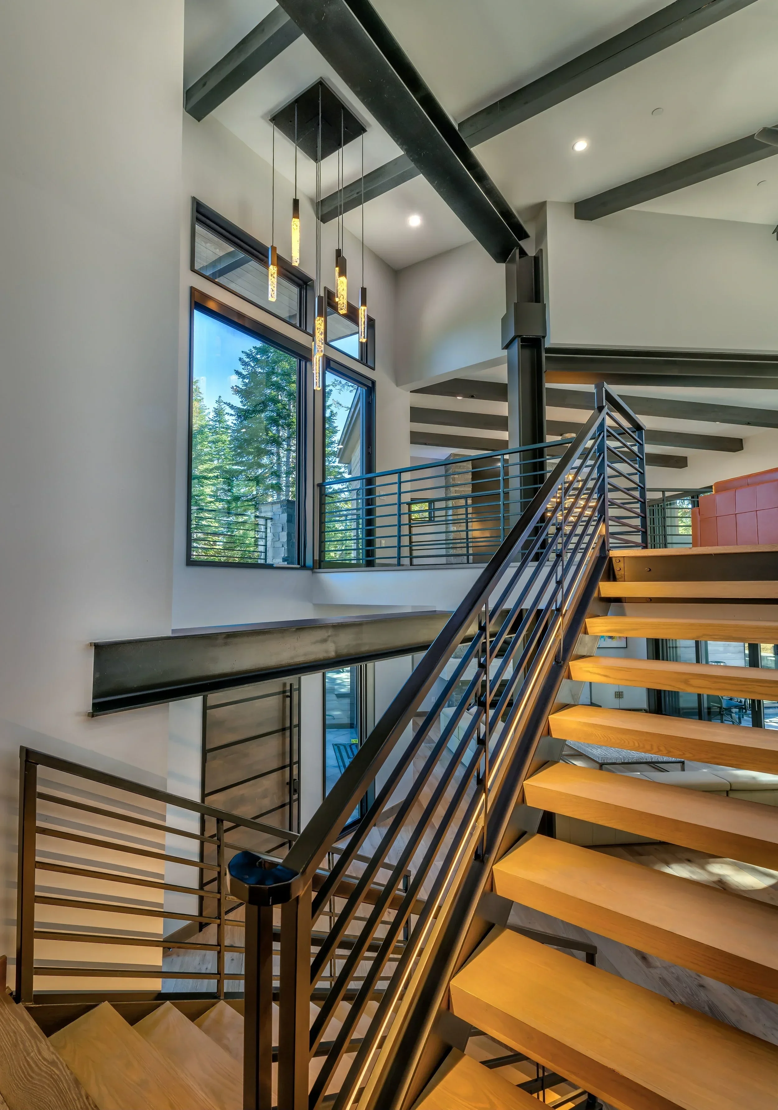 Interior view of a modern building with a staircase featuring wooden steps and black metal railings. Large windows reveal green trees outside. Modern pendant lights hang from the ceiling, and exposed beams are visible.