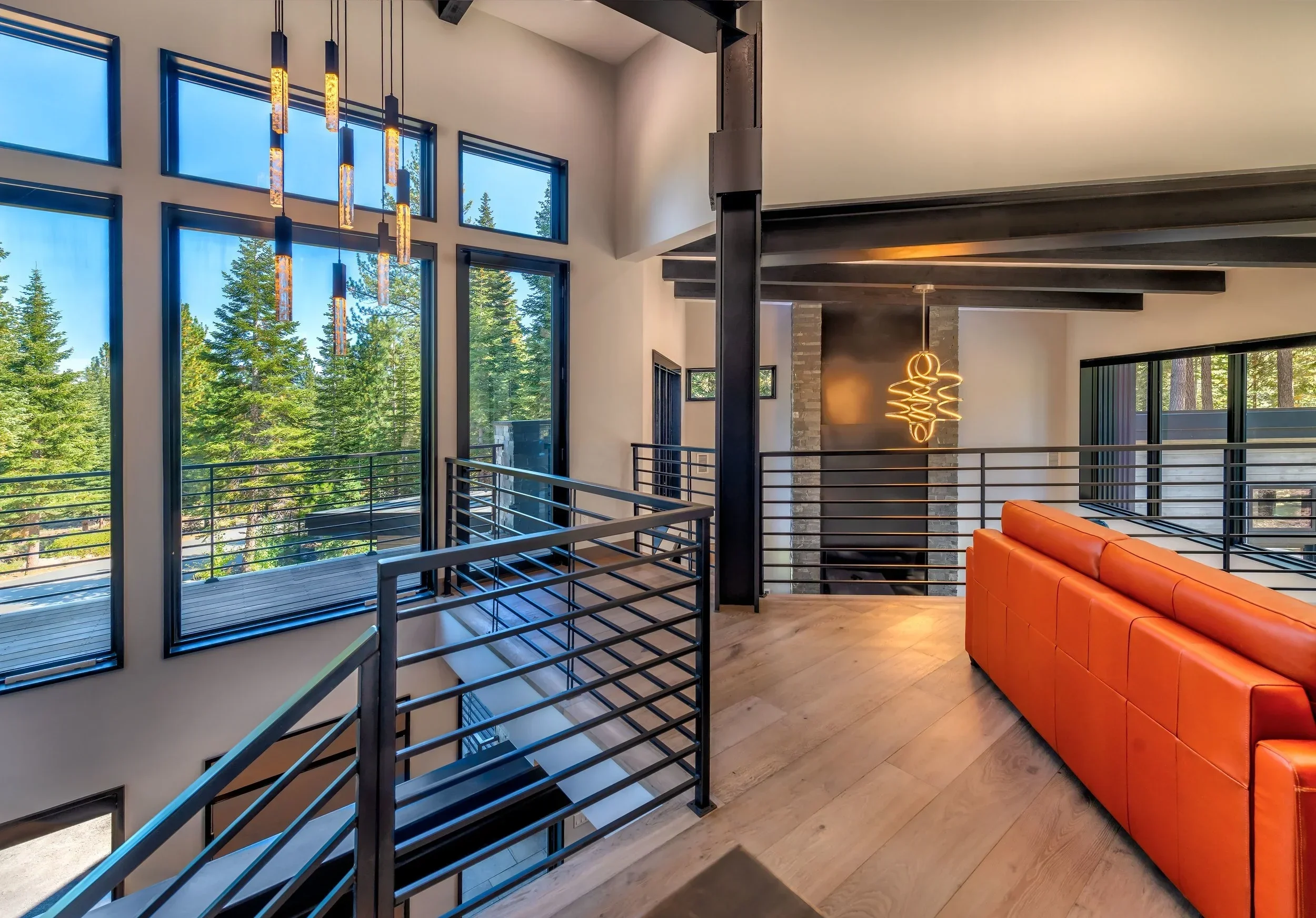 Modern living room with large windows overlooking a wooded outdoor area, featuring a black metal railing, a bright orange leather sofa, wooden flooring, and stylish lighting fixtures, including a pendant light and a wall-mounted sculpture light.