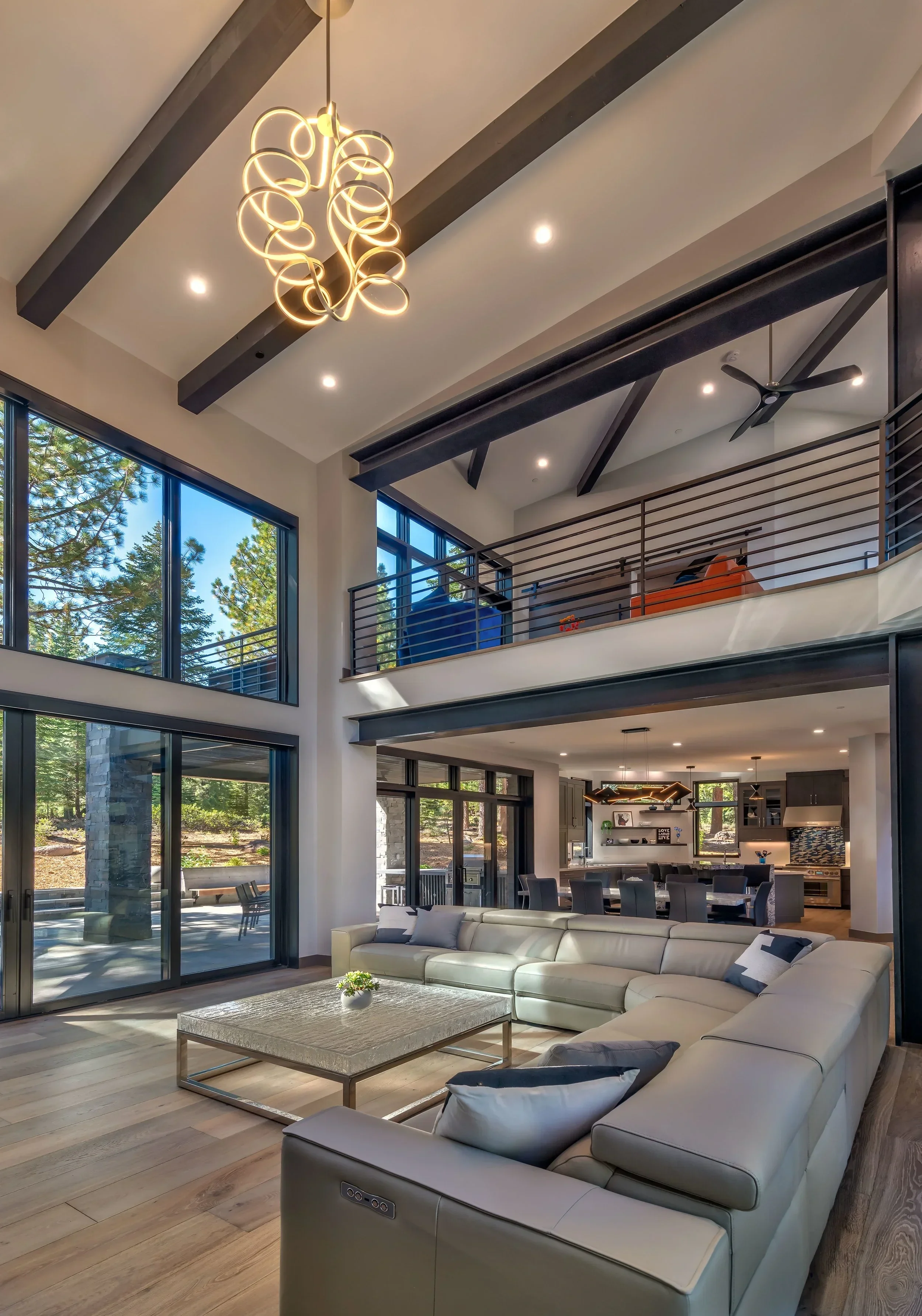 Interior view of a spacious modern living room with large floor-to-ceiling windows, a white sectional sofa with pillows, a wooden coffee table with a small plant, and a view of the outdoor patio and forest.