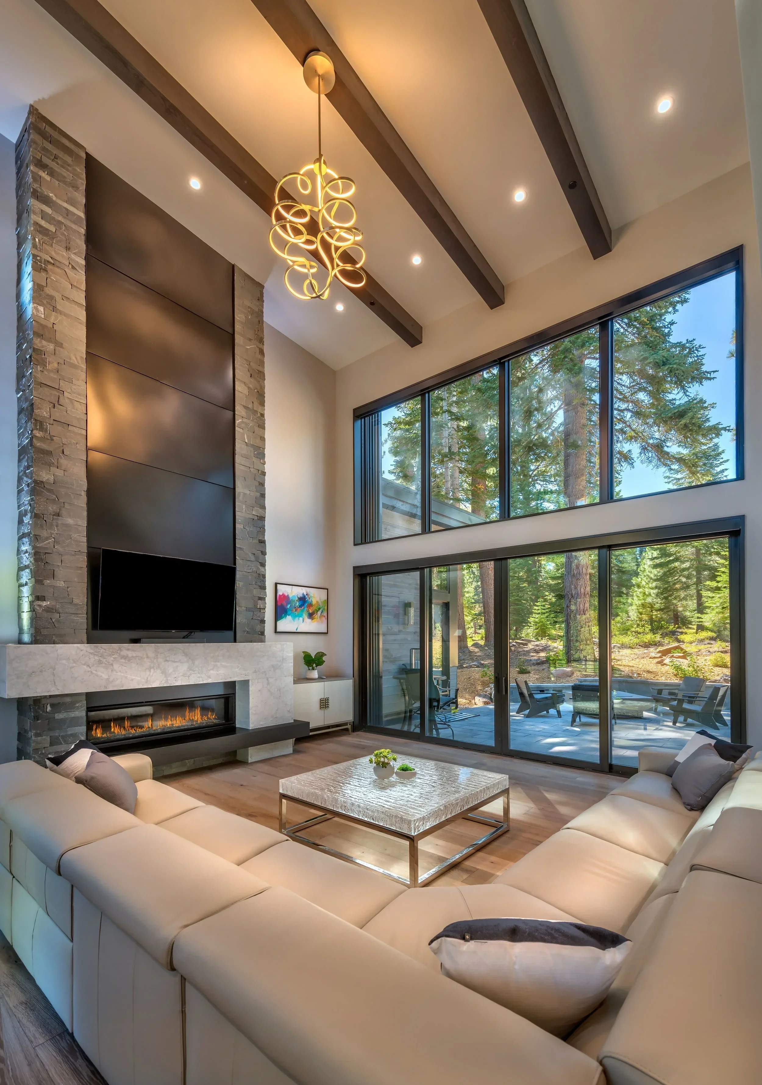 Living room with high ceiling, large floor-to-ceiling windows showing a wooded outdoor area, modern fireplace, beige sectional sofa, and contemporary chandelier.