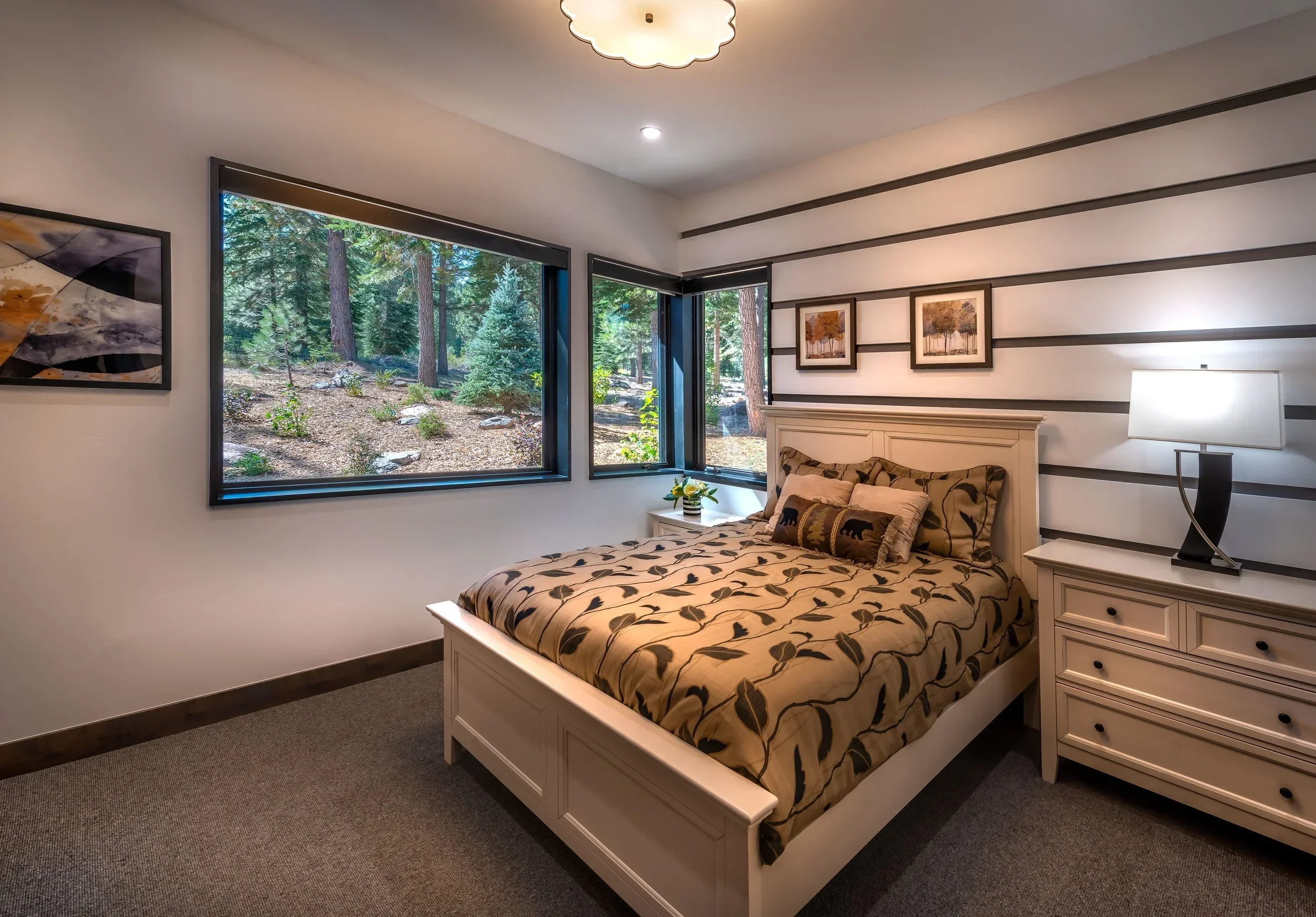 A bedroom with a white bed and a pillow with bear designs, a white nightstand with a lamp, and wall artwork. Large windows reveal a forest outside.