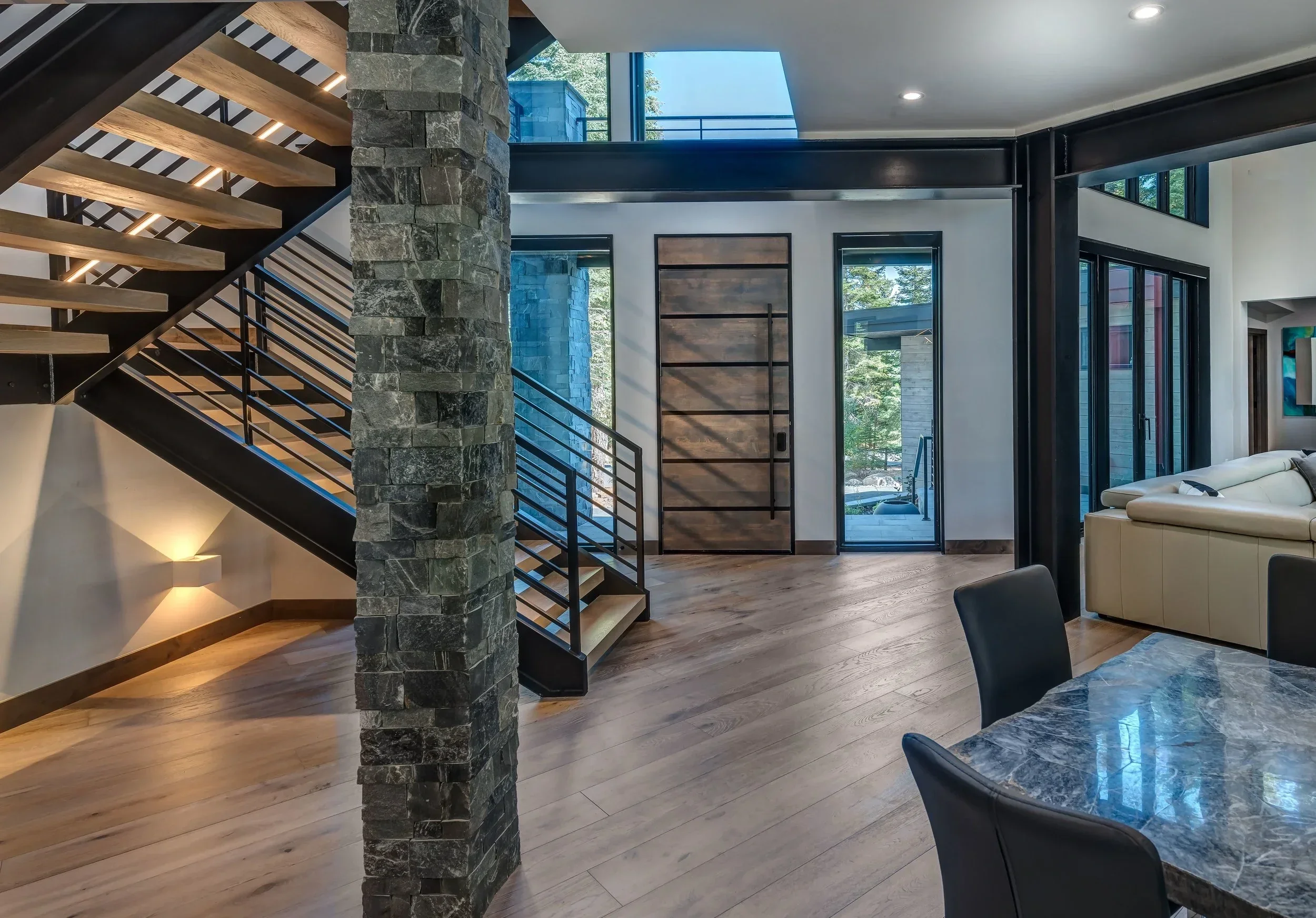 Modern interior of a house featuring a staircase with wooden steps, black metal railing, stone pillar, large windows, and a seating area with a white sofa and a marble dining table with black chairs.