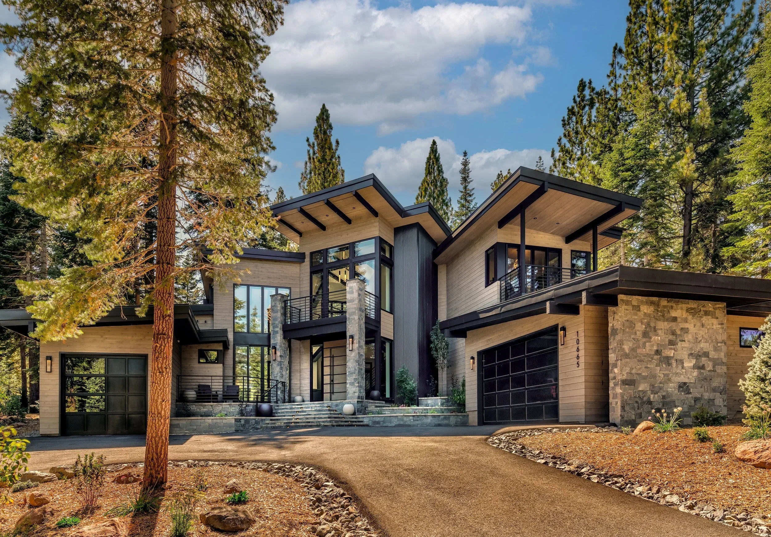 Modern multi-story house with large windows and a driveway surrounded by trees and landscaping.