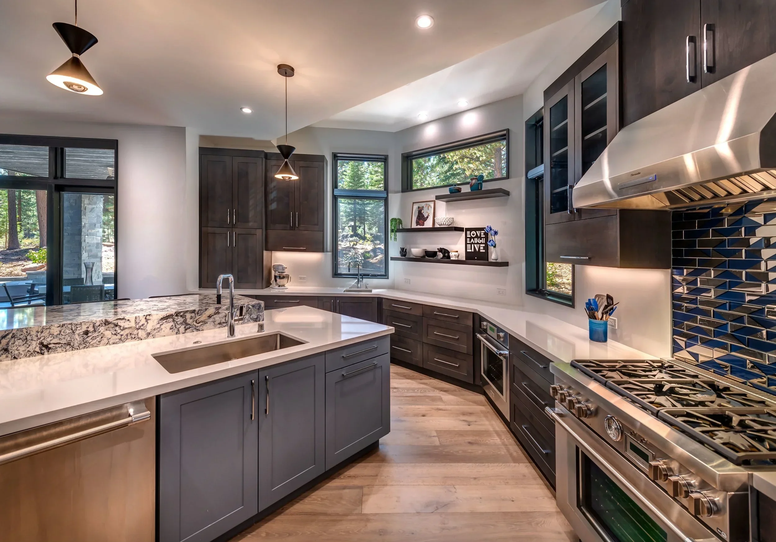 Modern kitchen with dark cabinets, white countertops, and stainless steel appliances. There is a kitchen island with a sink, open shelving on the wall, and large windows showing a wooded outdoor view.