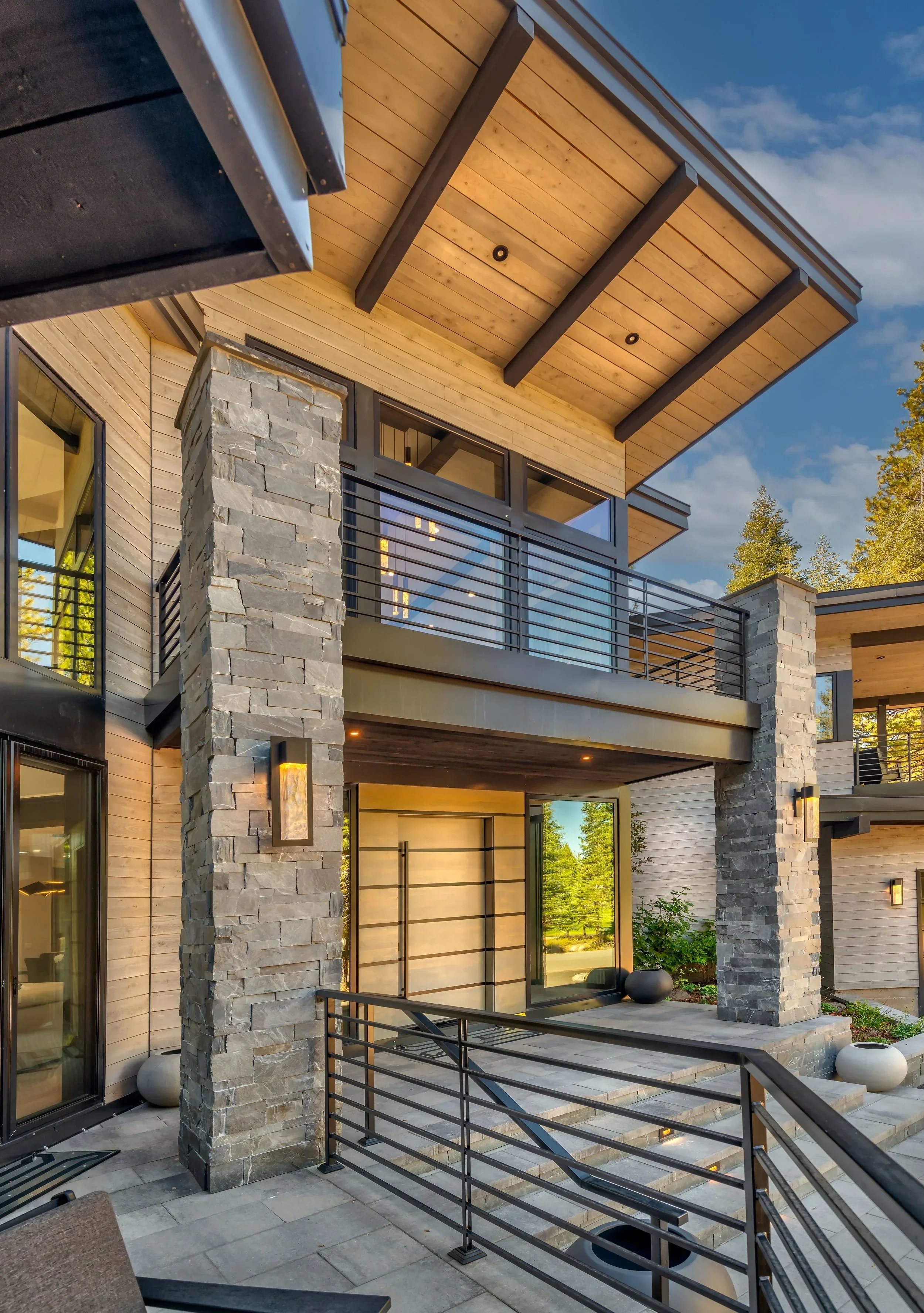 Exterior of a modern house with stone and wood siding, metal railings, and large glass windows under a wooden overhang, with outdoor seating and potted plants.