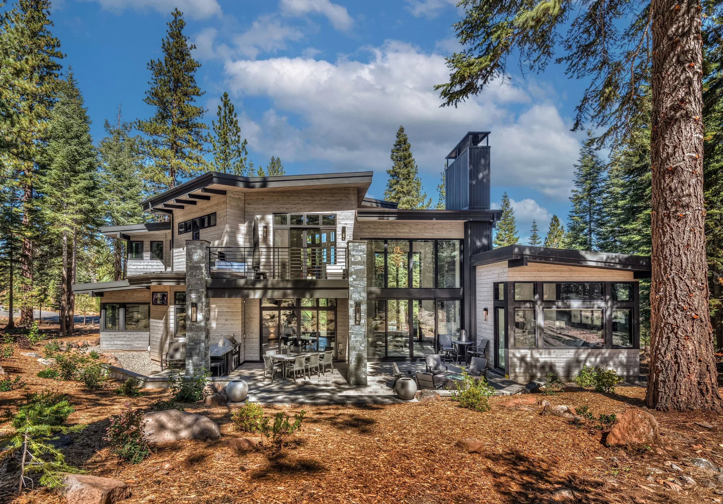 A modern, multi-story house with large windows, situated in a wooded forest, featuring a balcony and outdoor seating area.