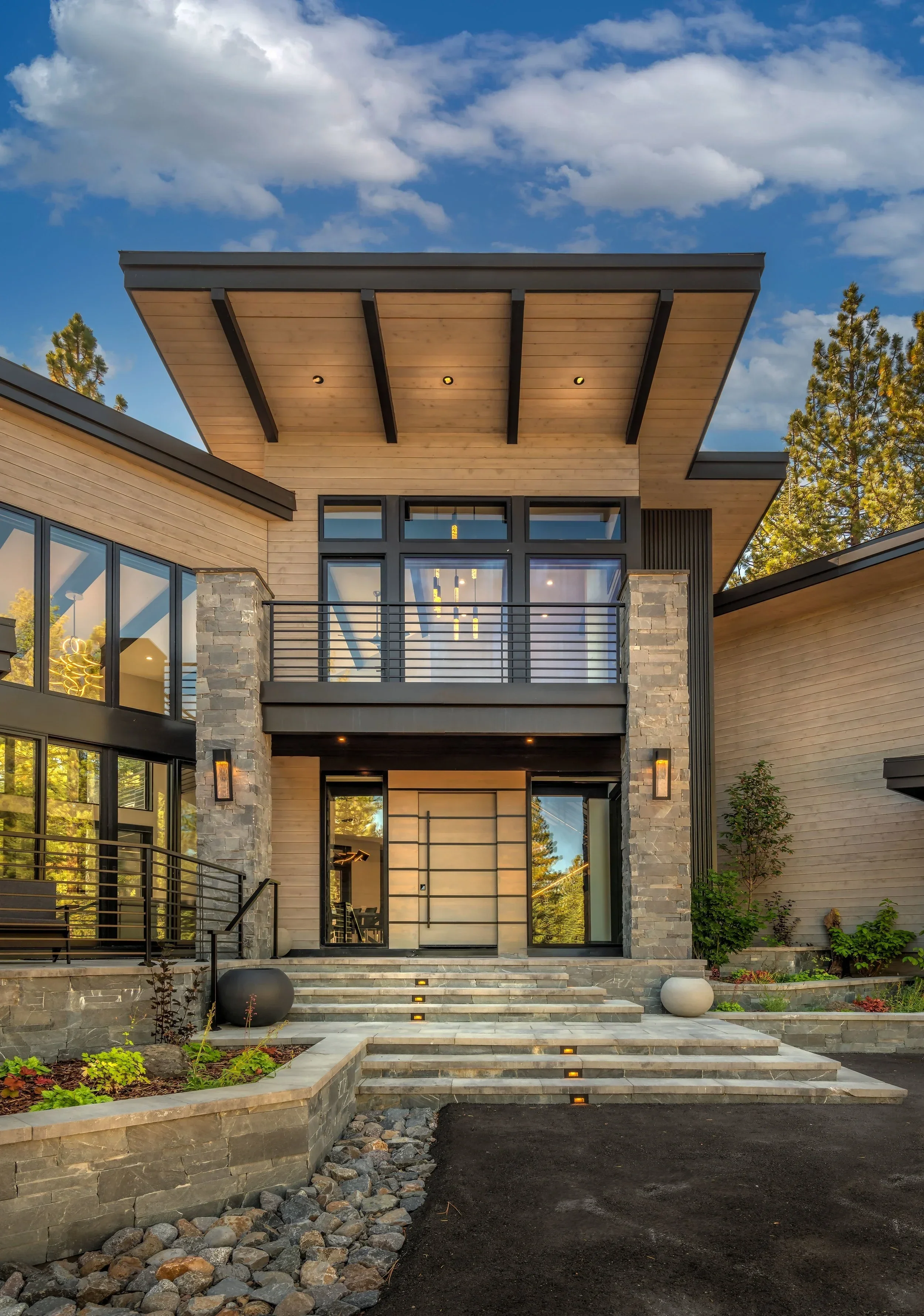 Modern house with stone and wood exterior, glass doors and windows, front steps with small lights, landscaped garden and trees, under a partly cloudy sky.