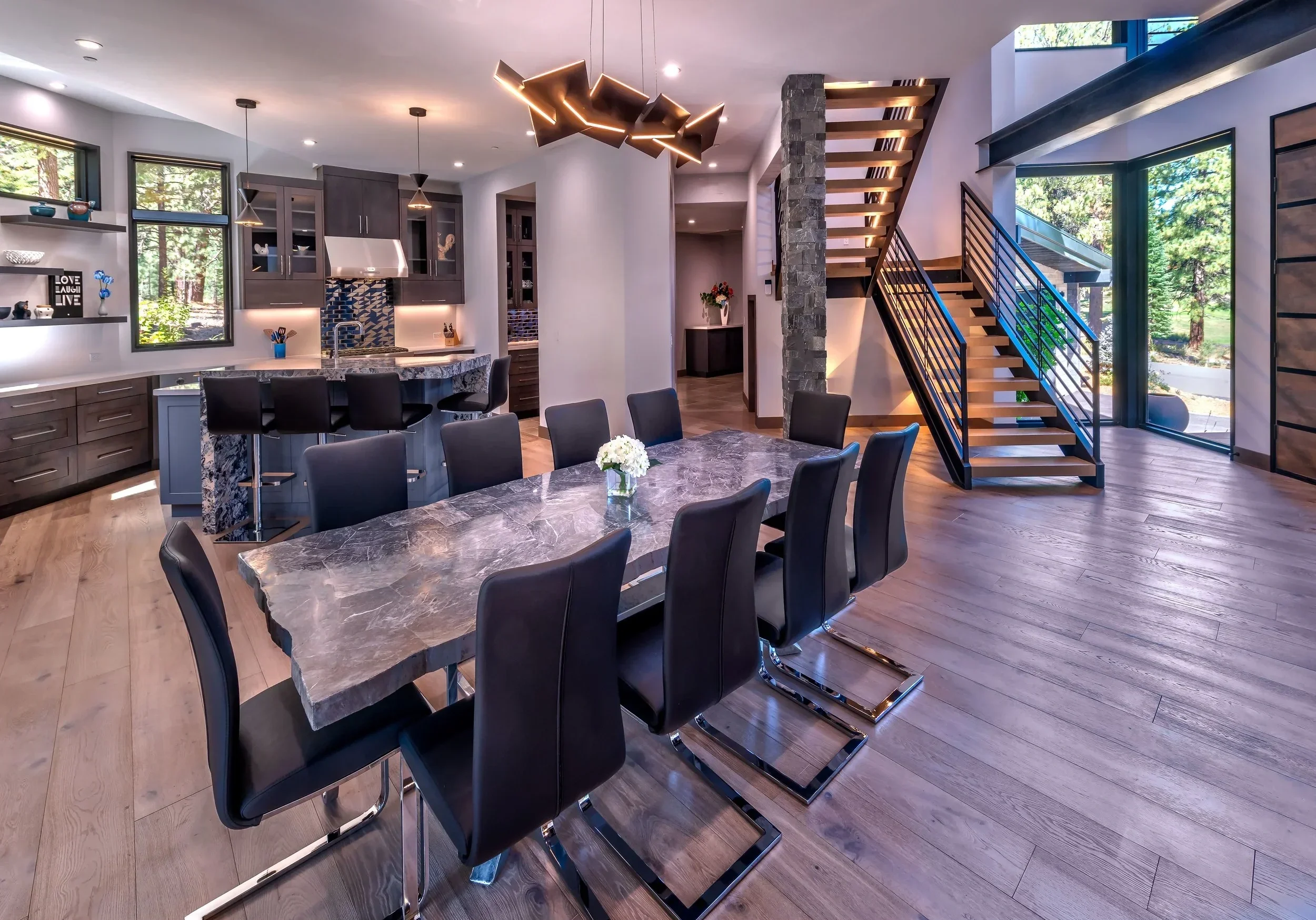 Modern open-concept kitchen and dining area with a large marble dining table, black chairs, a kitchen island with bar seats, dark cabinetry, a staircase with wood steps and black metal railing, large windows showing green trees outside, and contemporary light fixtures.