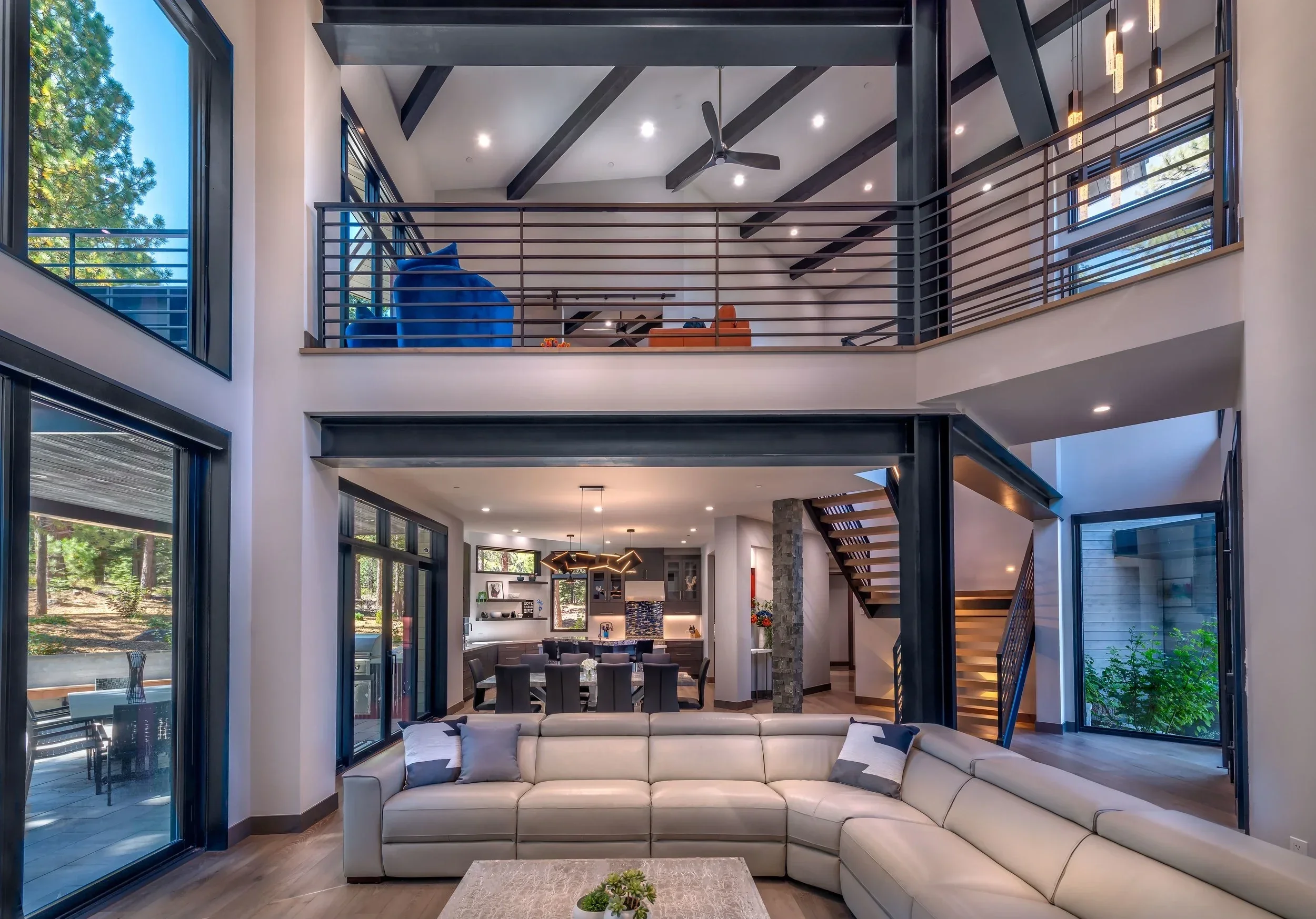 Spacious modern living room with a large white sectional sofa, floor-to-ceiling windows, and a view of the dining area and kitchen in the background. Two stories with open space, metal railings, and a high vaulted ceiling with black beams and a ceili