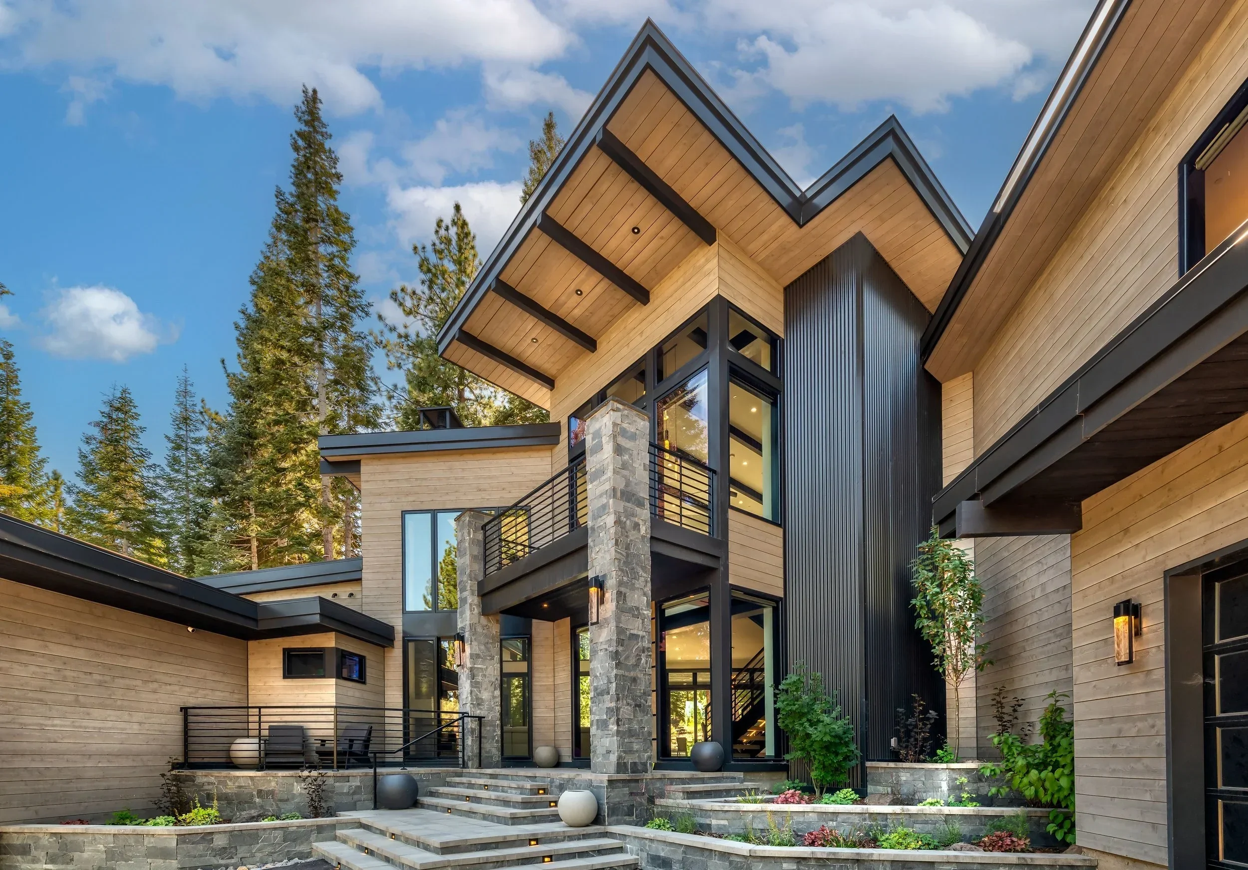 Modern house with large windows, wooden siding, stone pillars, and surrounded by tall trees under a partly cloudy sky.