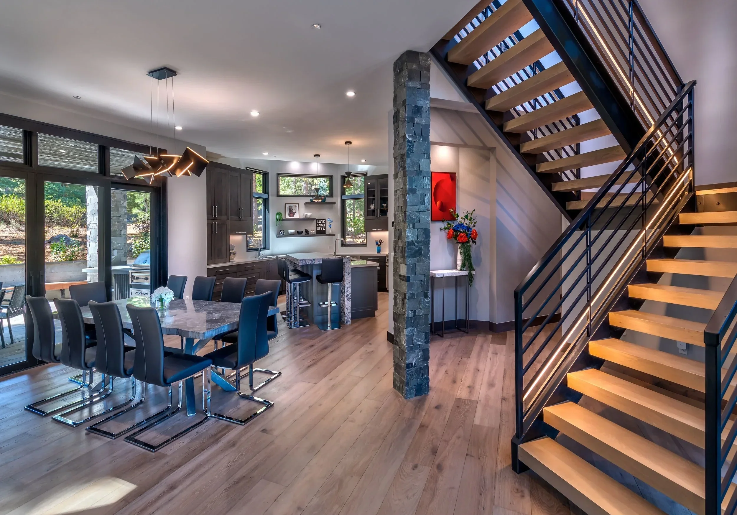 Open-concept living space featuring a dining area with a marble table and black chairs, a modern kitchen with gray cabinetry and a breakfast bar, a staircase with wooden steps and black metal railing, and large windows with a view of greenery outside