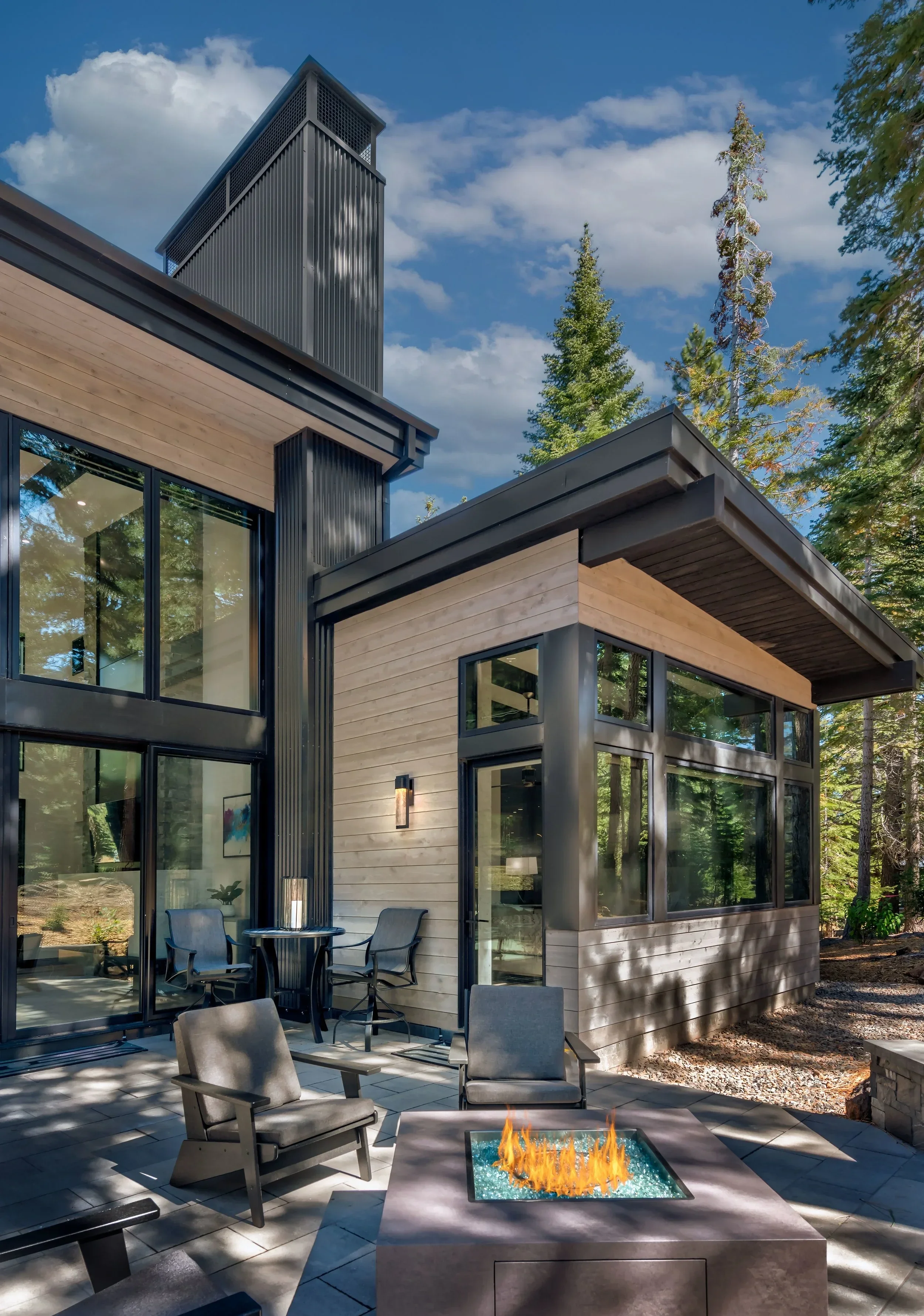 Modern house exterior with large glass windows, outdoor seating area with chairs and a fire pit, surrounded by tall trees under a partly cloudy sky.