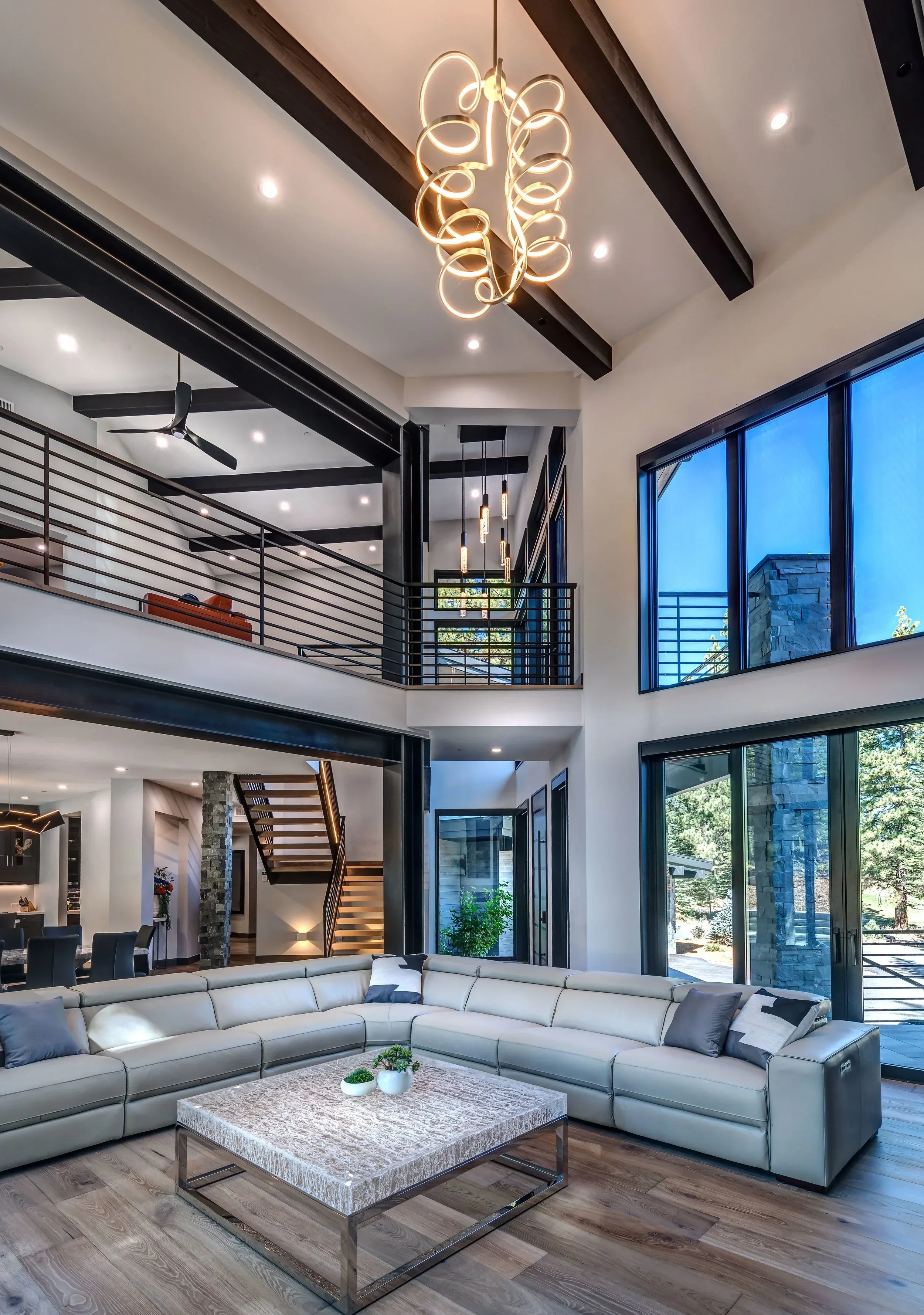 Spacious modern living room with high ceilings, large windows, and contemporary decor, including a white sectional sofa, a marble coffee table with small plants, and stylish lighting fixtures.