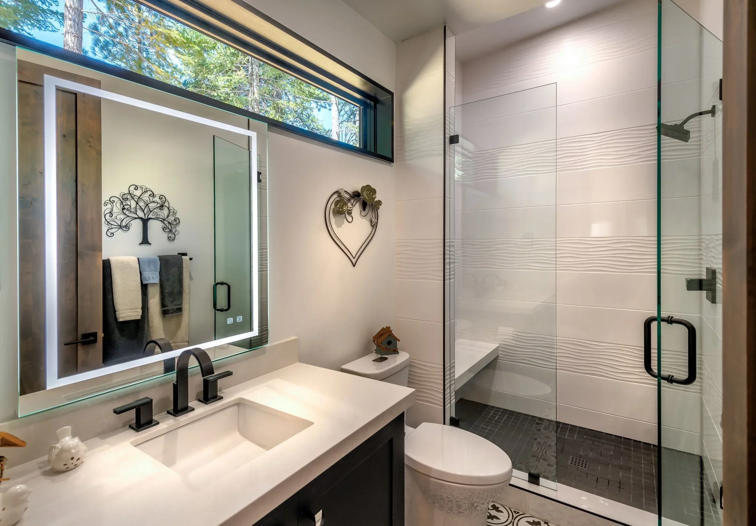 Modern bathroom with a large backlit mirror, white sink with black fixtures, a floating white toilet, and a glass-enclosed shower area with textured white tiles and a black tiled floor. Decor includes wall art and small decorative items on the counte