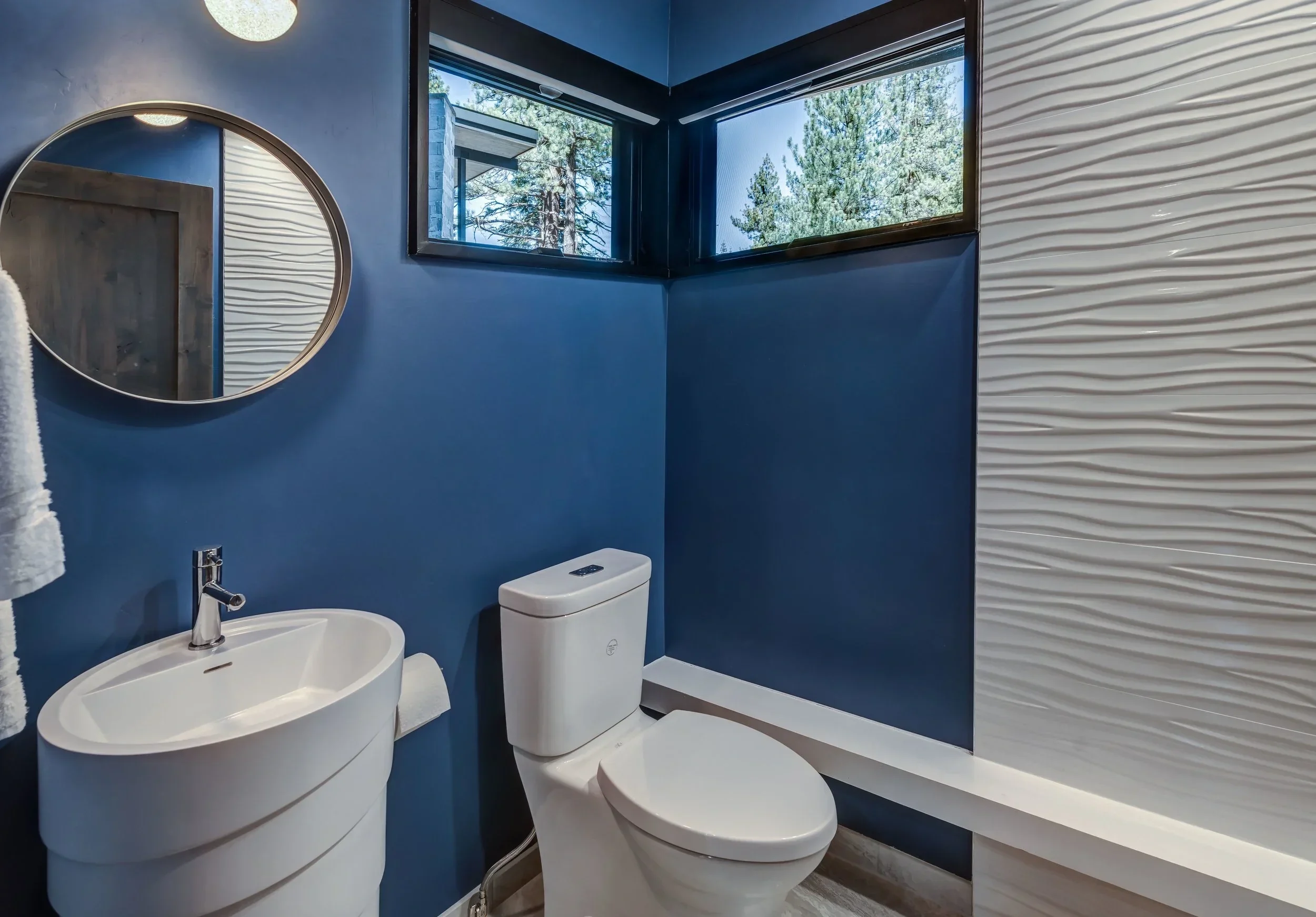 Modern bathroom with blue walls, a white sink with curved edges, a toilet, a round mirror, and a window showing trees outside.