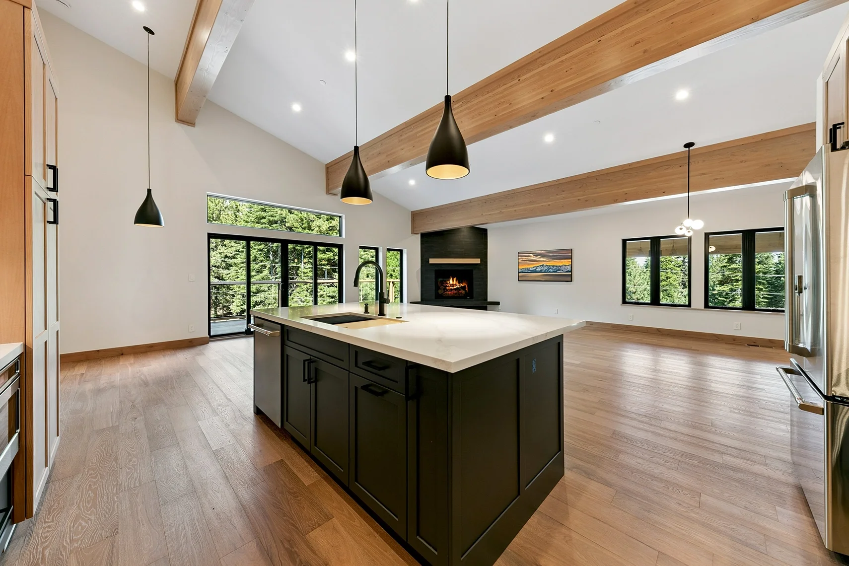 Open-concept kitchen and living area with a large island, pendant lighting, natural wood flooring, white walls, exposed ceiling beams, and abundant windows and doors.