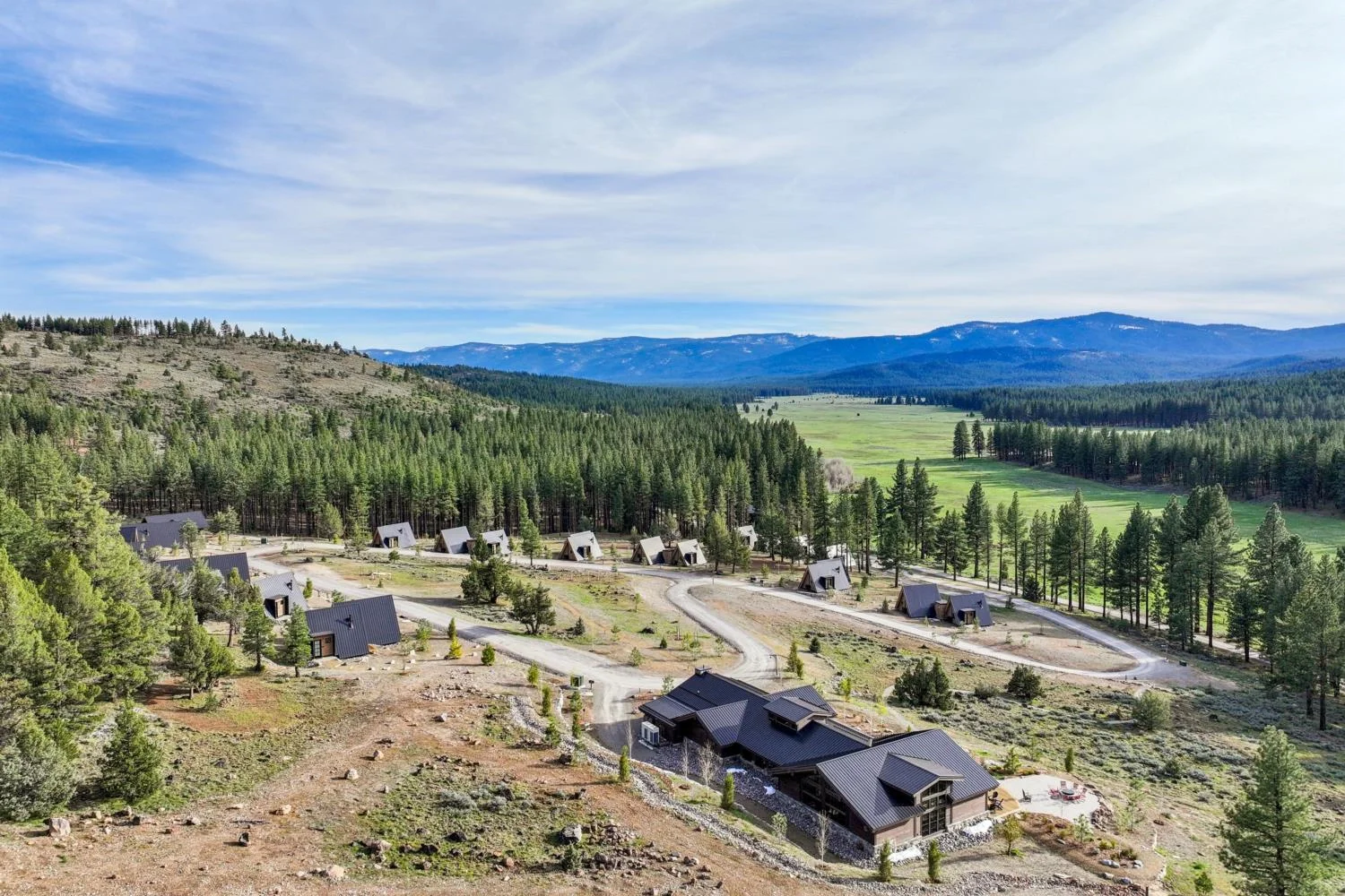 Aerial view of A-frame cabins and modern mountain buildings along a winding hillside road overlooking a pine forest, open valley, and distant mountains.