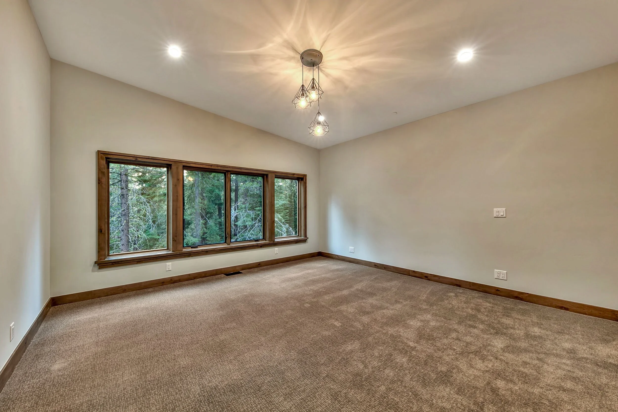 Empty living room with beige carpet, beige walls, a large window with natural wood trim, and a modern chandelier with exposed bulbs hanging from the ceiling.