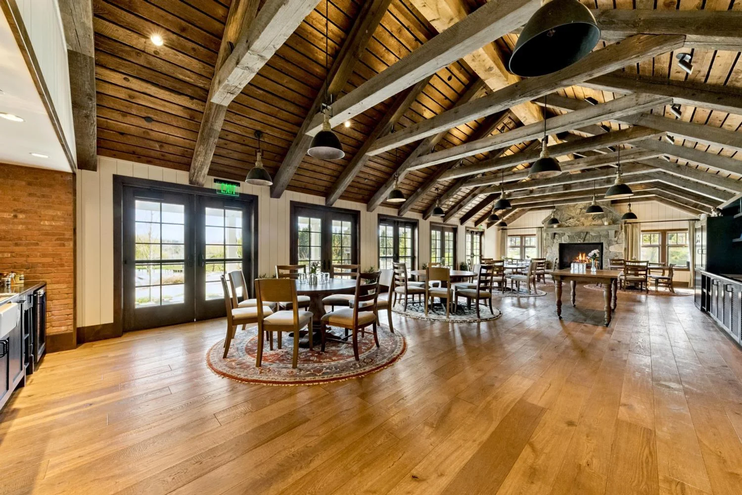 Large seminar room with round tables, a vaulted wood ceiling, exposed timber trusses, black pendant lights, glass doors, and a stone fireplace.