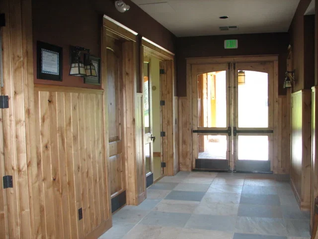 Interior of a building with wooden paneled walls, glass double doors leading outside, and a tiled floor.