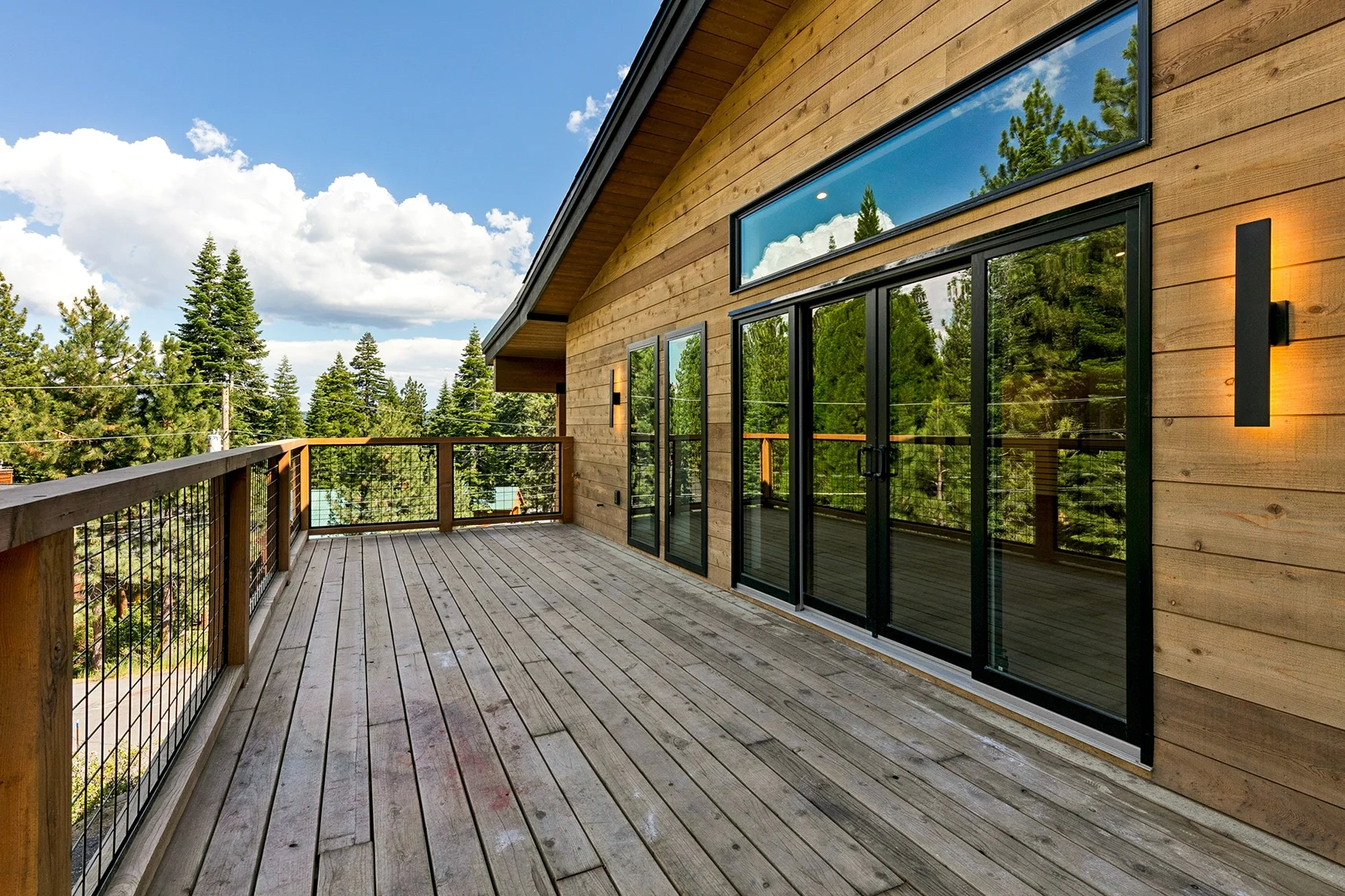 Modern outdoor deck with natural wood siding, six glass window and sliding door panels, surrounded by a forest landscape under a blue sky with white clouds.