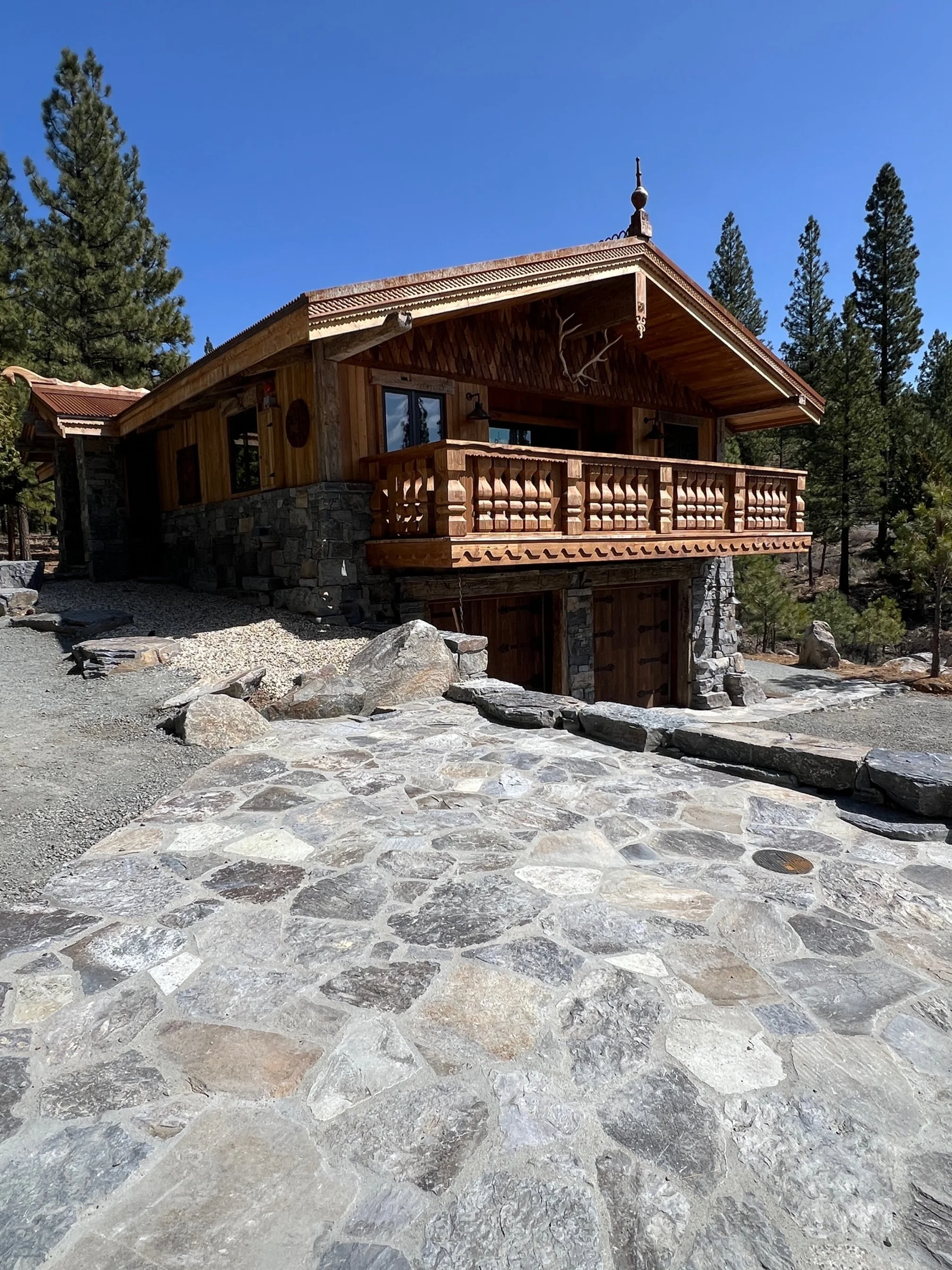 A rustic wooden house with a stone foundation and a large elevated balcony, surrounded by tall pine trees, under a clear blue sky.