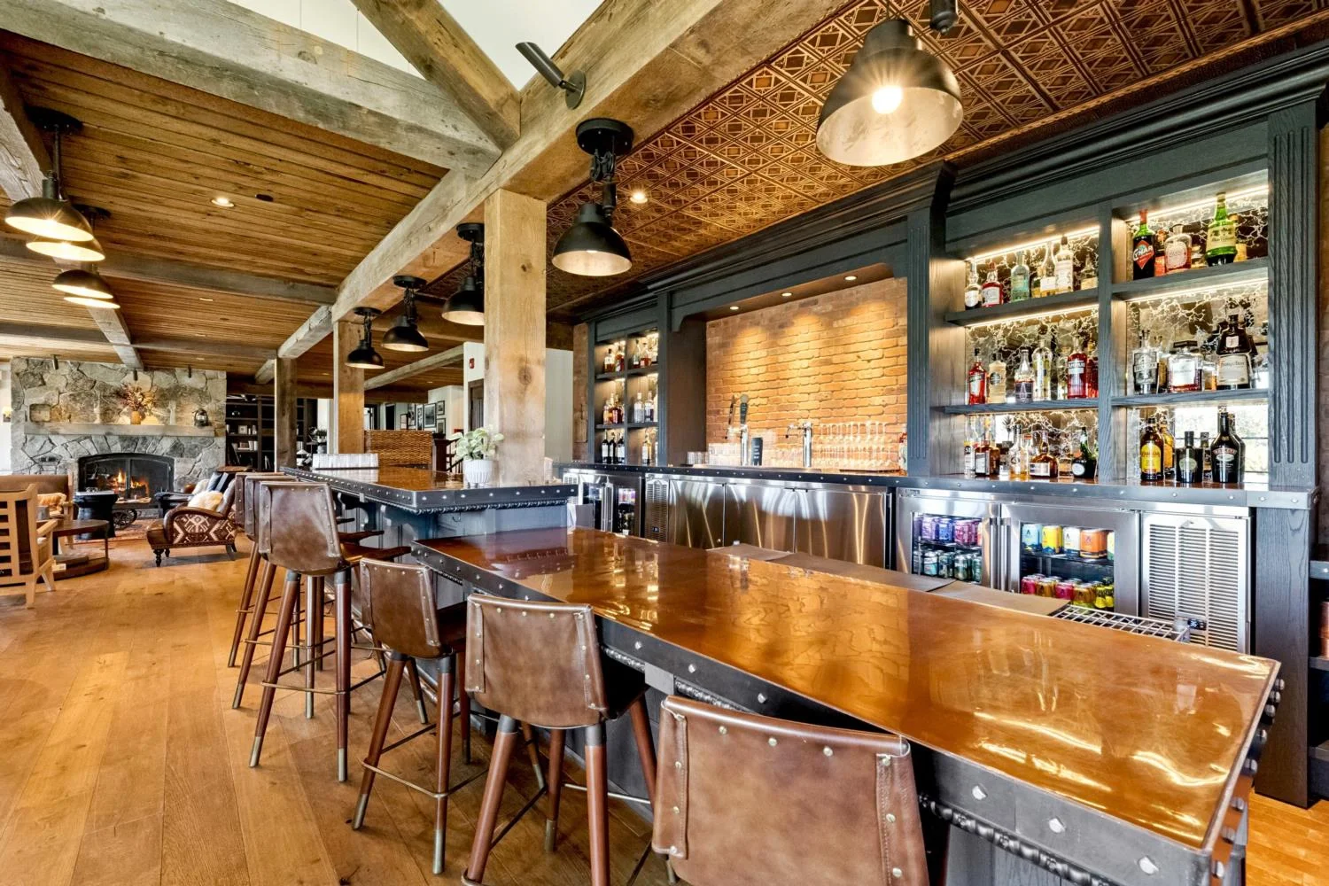 intage Western-style lodge bar with a pressed copper ceiling, polished wood counter, leather stools, illuminated shelving, brick backsplash, and exposed timber beams.