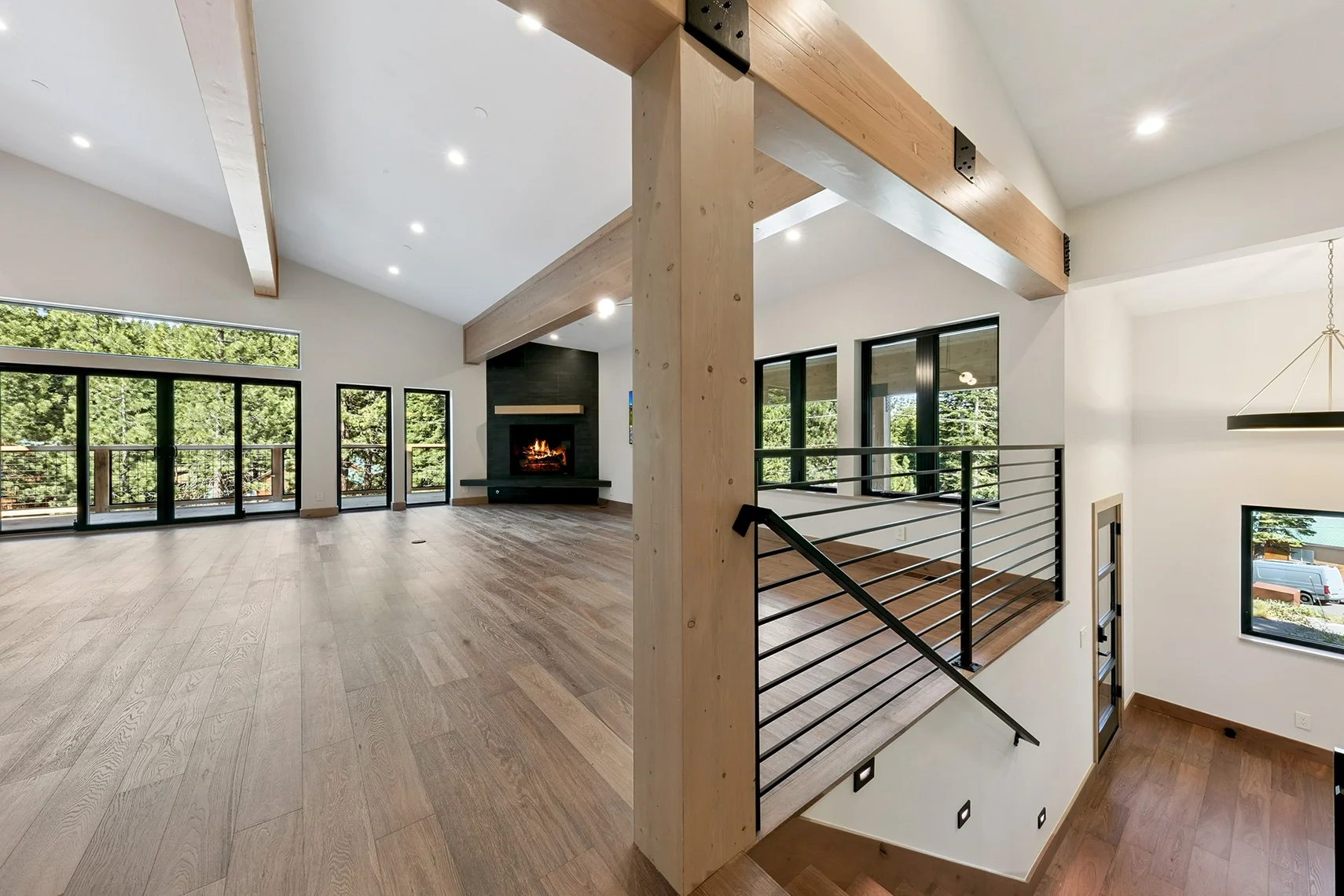 Open-concept modern mountain living area featuring hardwood floors, a stone fireplace hearth, horizontal iron stair railings, and expansive sliding glass doors opening to the outdoors.