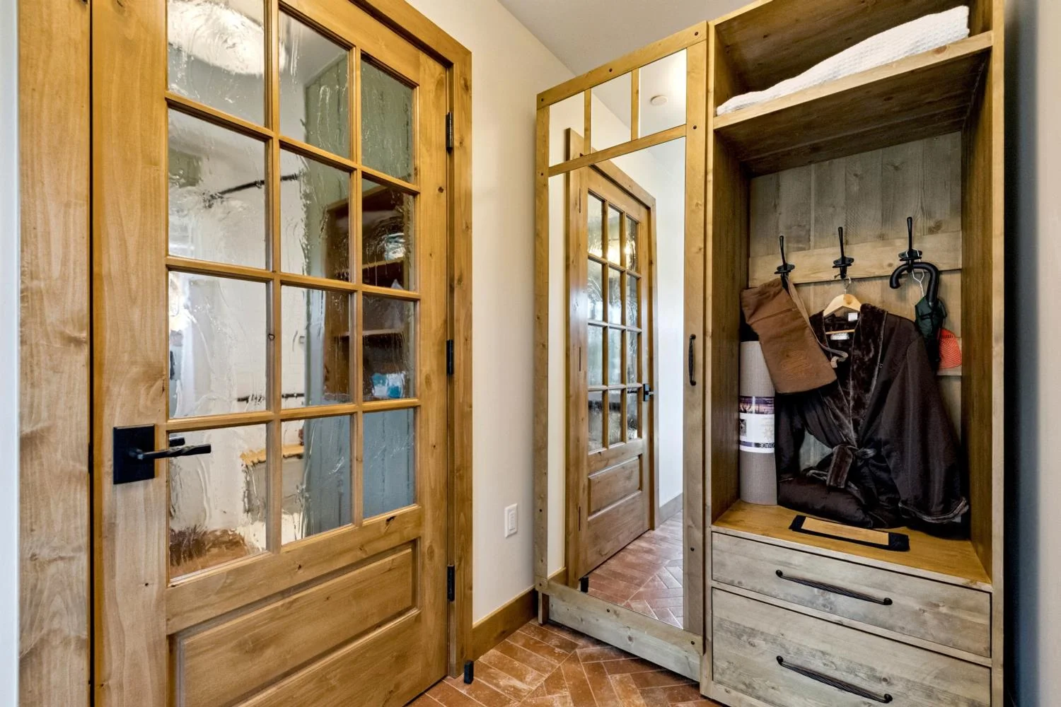 Private cabin entry with a built-in wood wardrobe, full-length mirror, textured glass door, black hardware, and herringbone brick tile flooring.