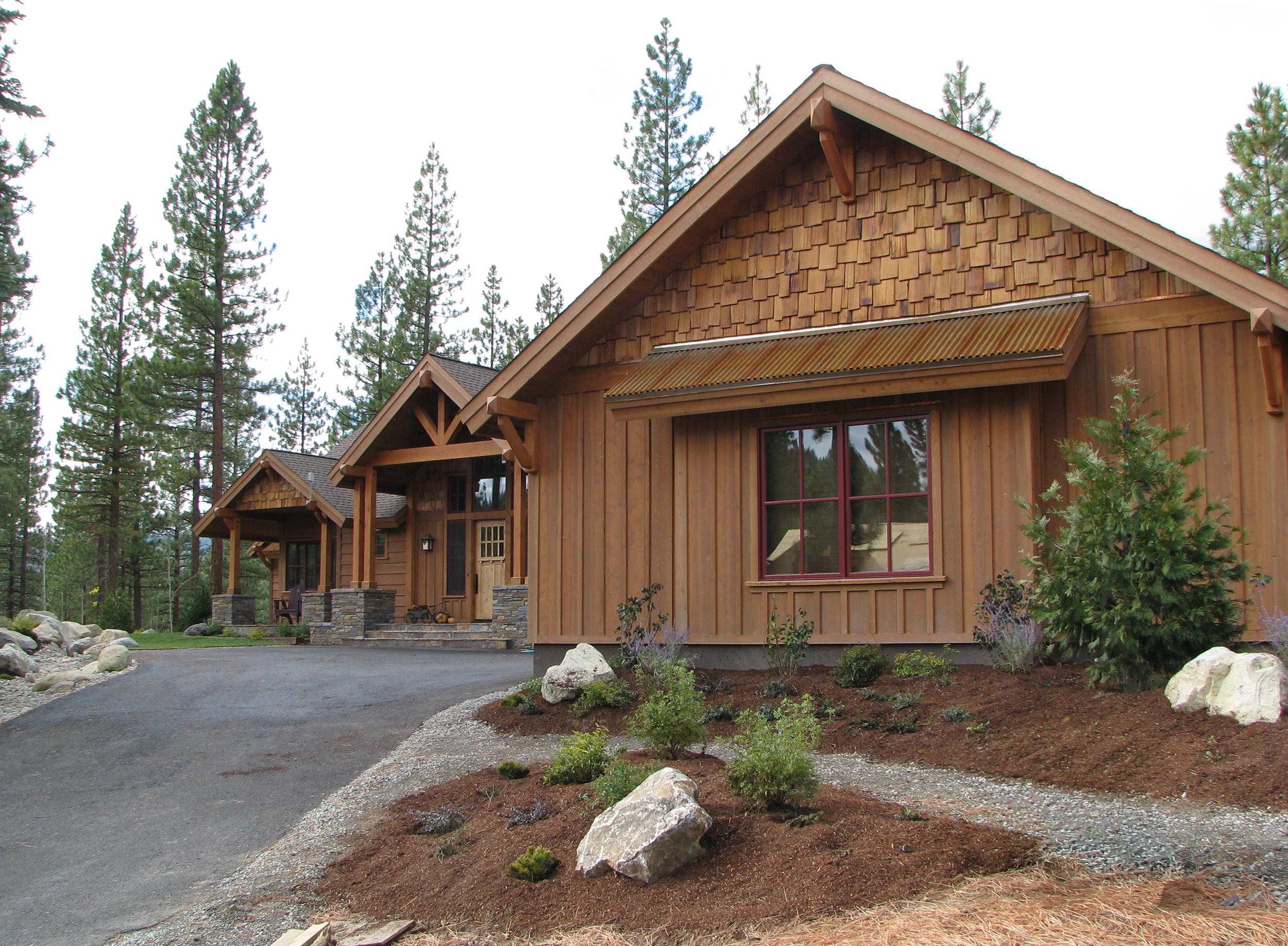 Single level home with brown wood and shingle siding, red trim and corrugated metal roof accents with new landscaping and pine trees in the background.