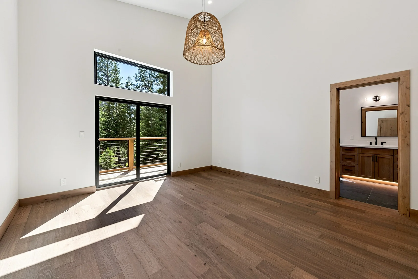 Modern bedroom with natural wood flooring, an attached bathroom, a rattan pendant light, and sliding glass doors opening to a private deck.
