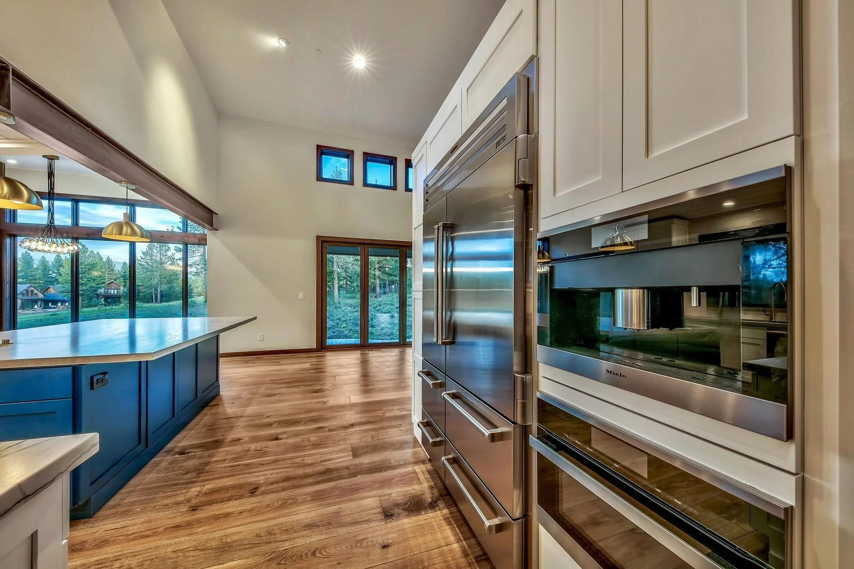 Modern kitchen with wooden floor, stainless steel refrigerator, built-in oven, and large windows overlooking greenery.