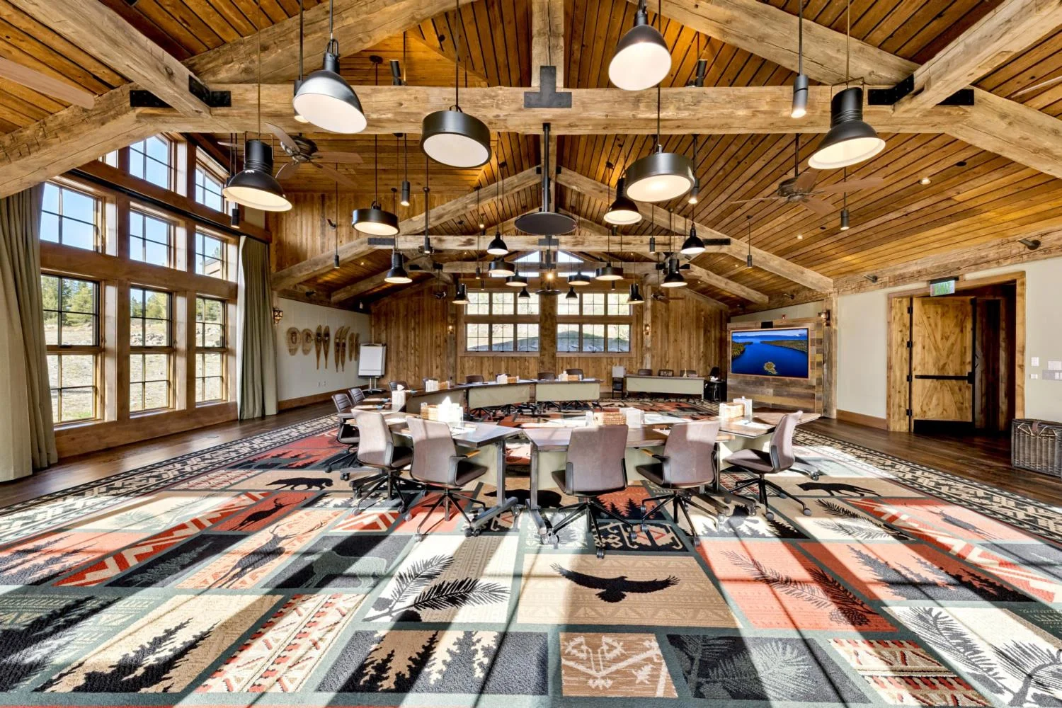 Large conference room with tables arranged in a circle, rolling chairs, a colorful nature-patterned carpet, vaulted wood ceiling, exposed timber trusses, varied pendant lights, and expansive windows.