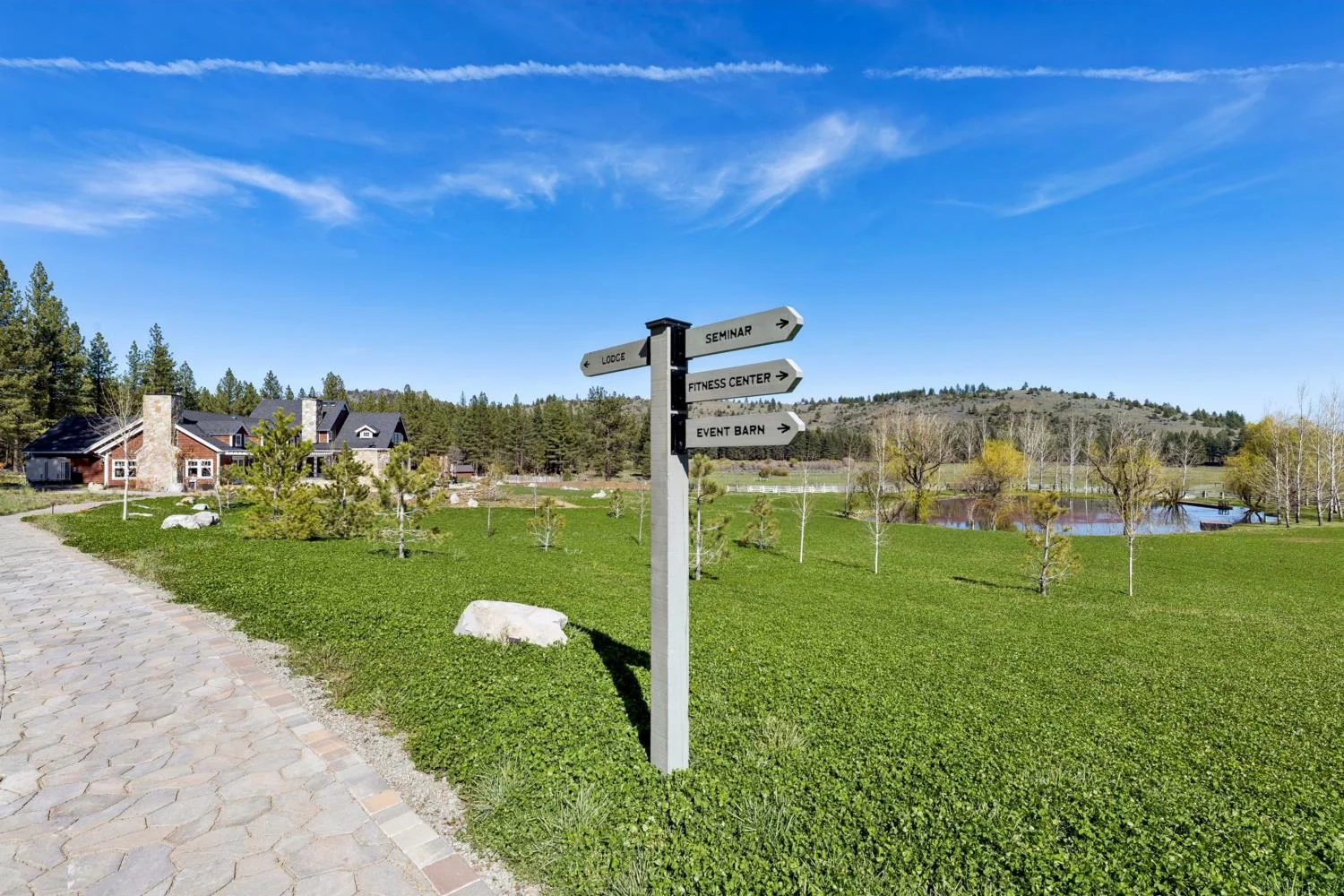 Directional sign for the lodge, seminar building, fitness center, and event barn beside a stone pathway overlooking a green meadow and pond beneath a blue sky.
