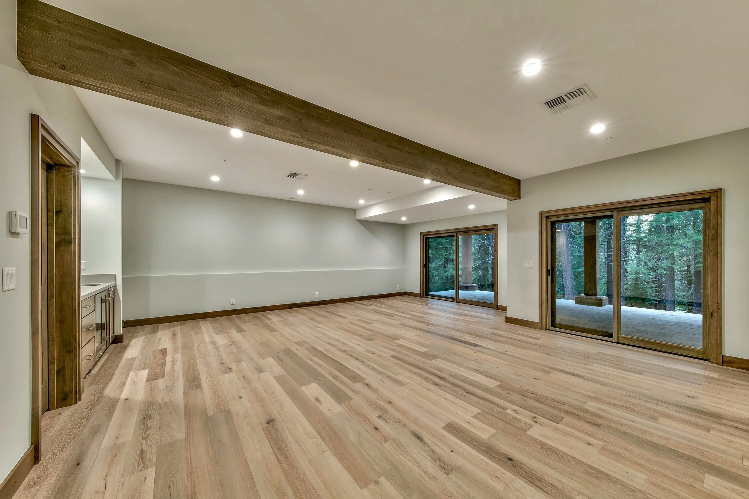 Empty living room with hardwood floors, a wooden beam on the ceiling, sliding glass doors opening to a wooded outdoor area, and recessed lighting.