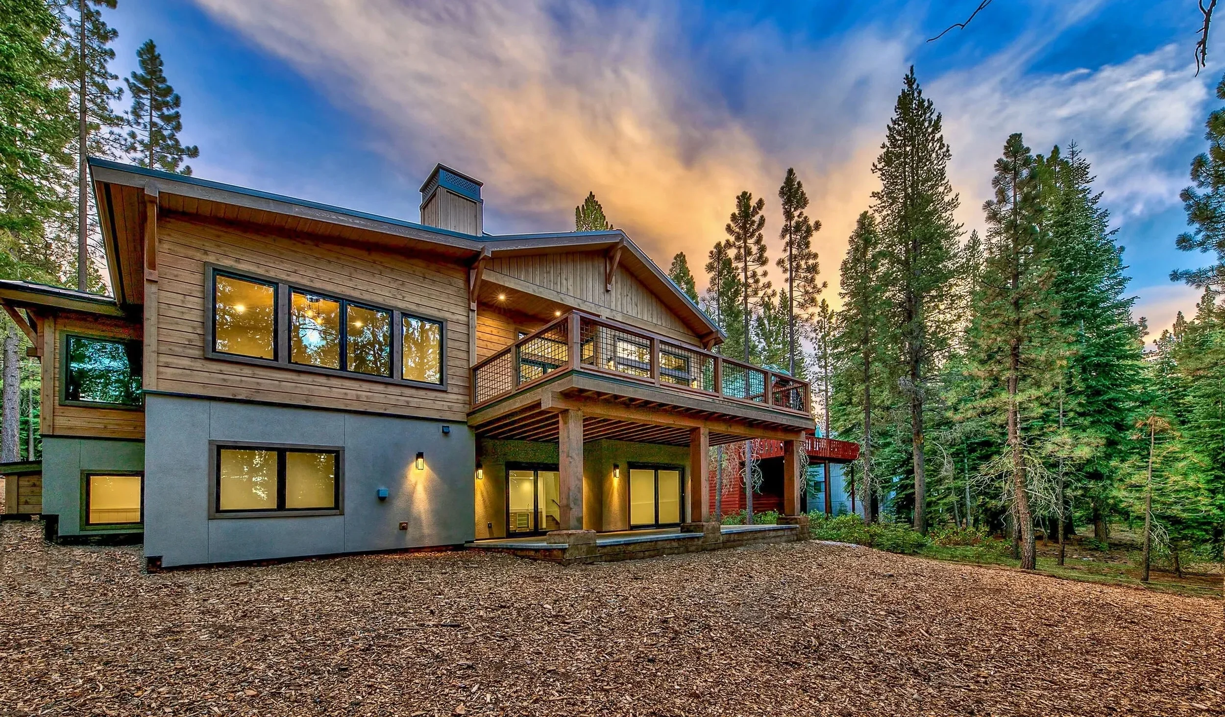 A modern house with wooden and concrete exterior, large windows, and a balcony surrounded by trees at sunset.