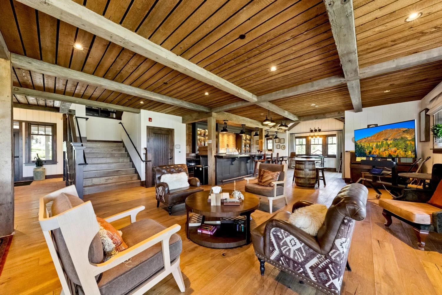 Large ranch-style lodge lounge with wood flooring, a plank ceiling, exposed beams, leather seating, a staircase, and a built-in bar.