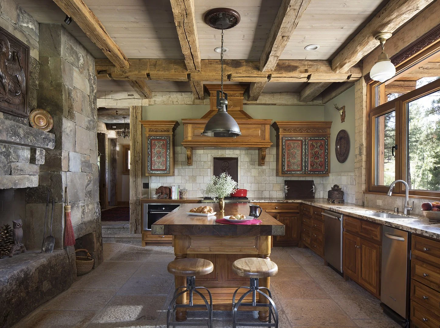 Rustic kitchen with wooden beams, stone fireplace, and natural light from large windows, featuring a kitchen island with two stools.