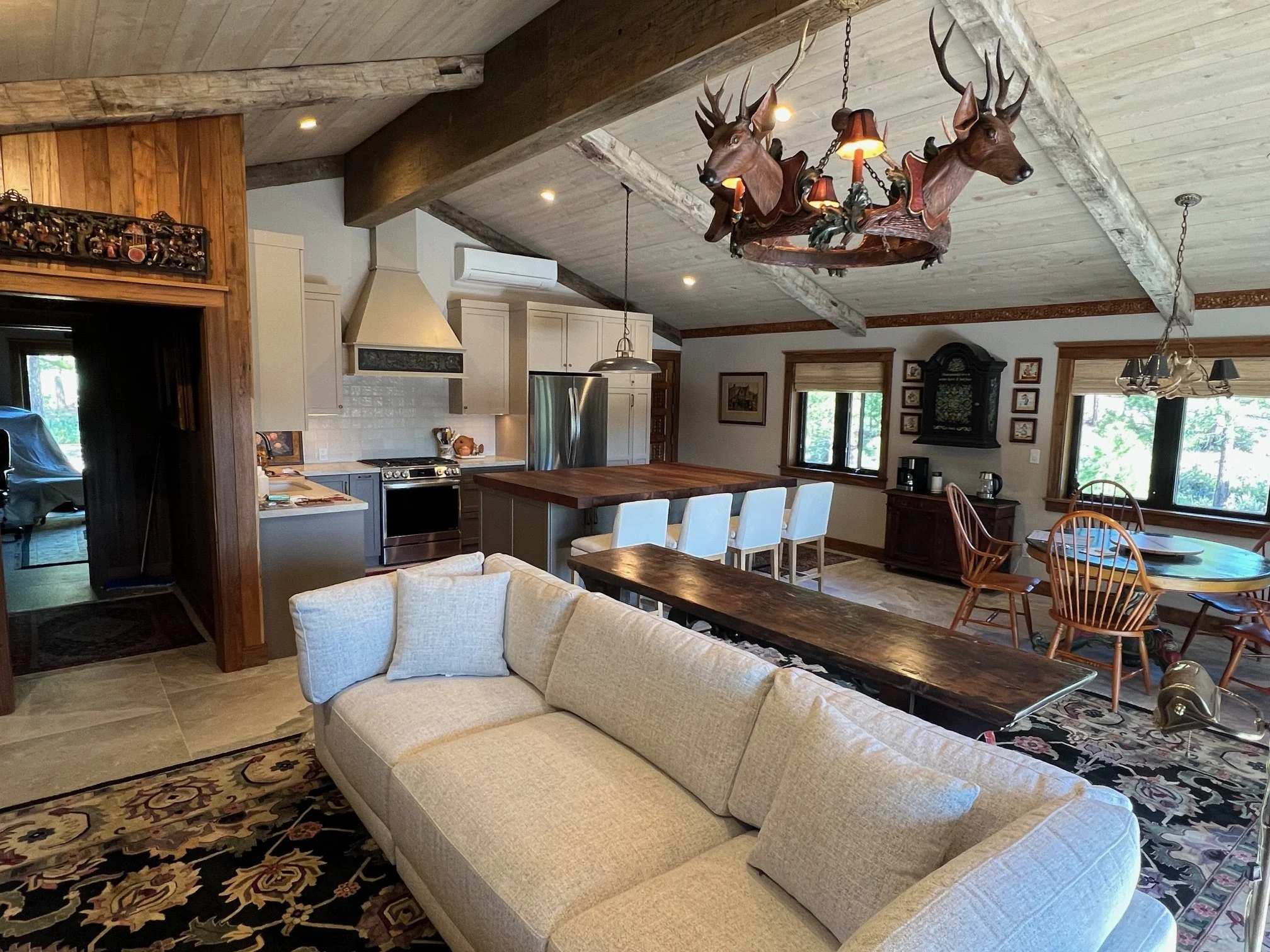 A rustic living room and kitchen area with wooden beams, a chandelier made of deer heads, a beige sofa, a round dining table with wooden chairs, and large windows showing a view of trees outside.