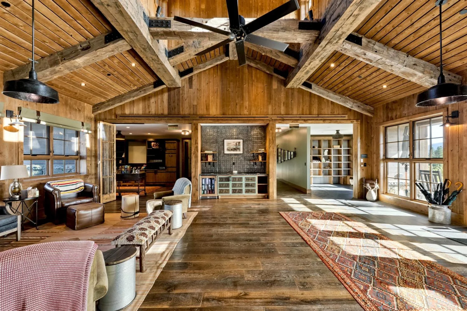 Spacious lodge lounge with dark wood flooring, a vaulted wood ceiling, exposed timber beams, a large overhead fan, leather seating, patterned rugs, a built-in refreshment station, and expansive windows.