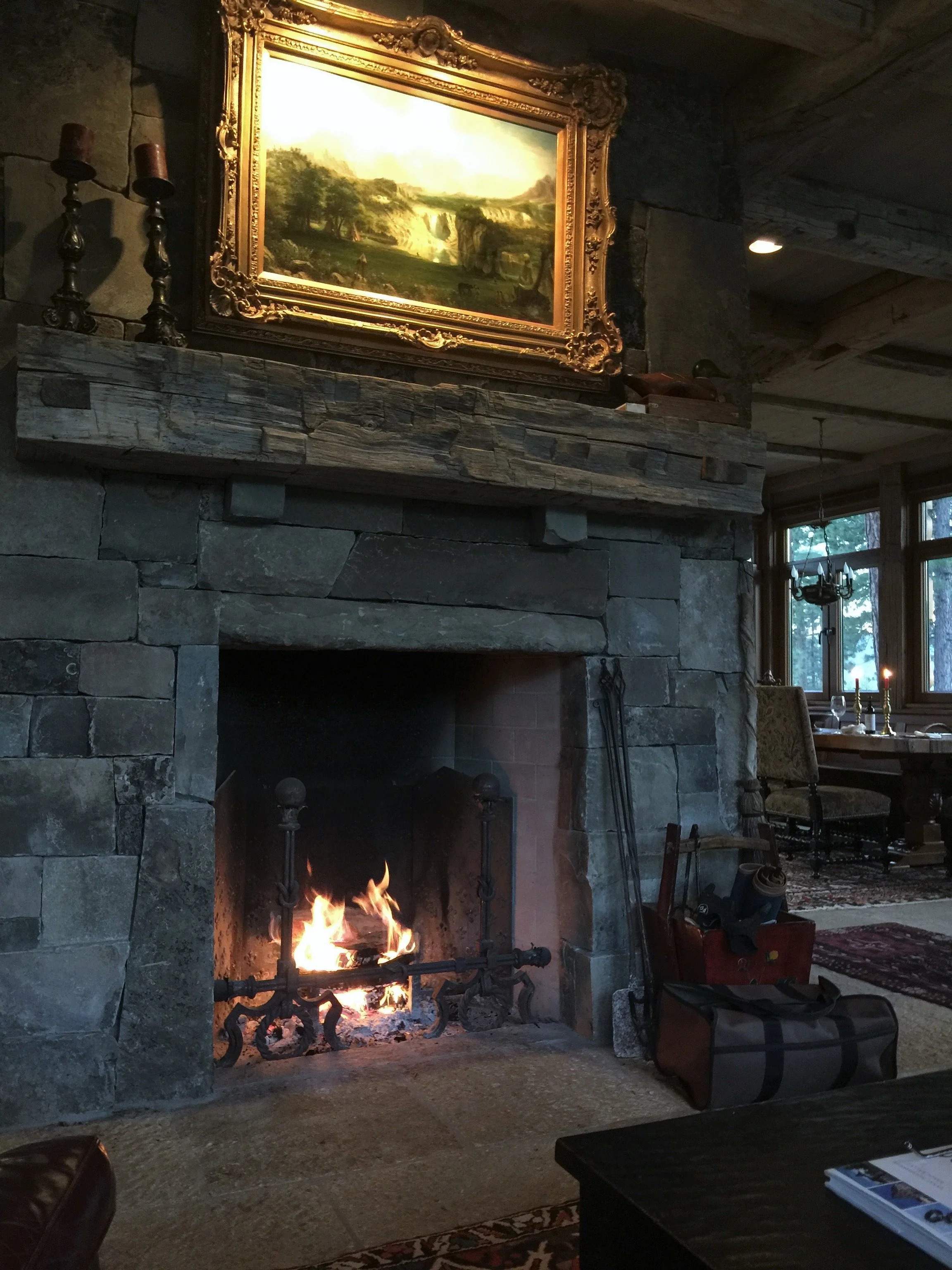 A cozy living room corner with a lit fireplace made of large stone bricks, topped with a rustic wooden mantel. Above the mantel, a framed landscape painting depicts a lush green scene with trees, waterfalls, and hills. To the right, there is a dining