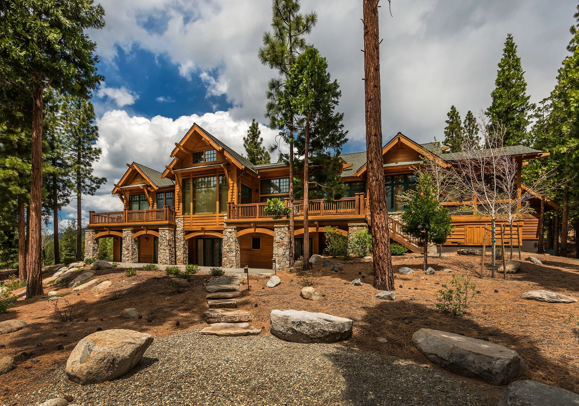 Large rustic mountain home in open wooded setting, with spacious decks, large windows and stone columns  with wooden arches between on lower level.