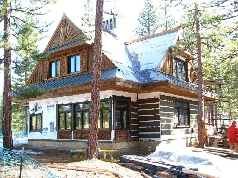 A two-story house under construction in a wooded area, with wooden framing and black window frames, surrounded by tall trees.