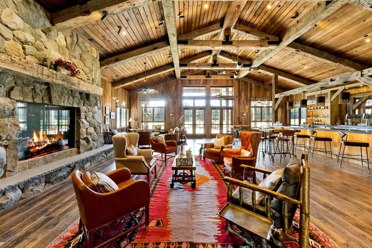 Large modern mountain lodge lounge with a floor-to-ceiling stone fireplace, vaulted wood ceiling, exposed timber trusses, leather seating, a colorful patterned rug, and a full bar.