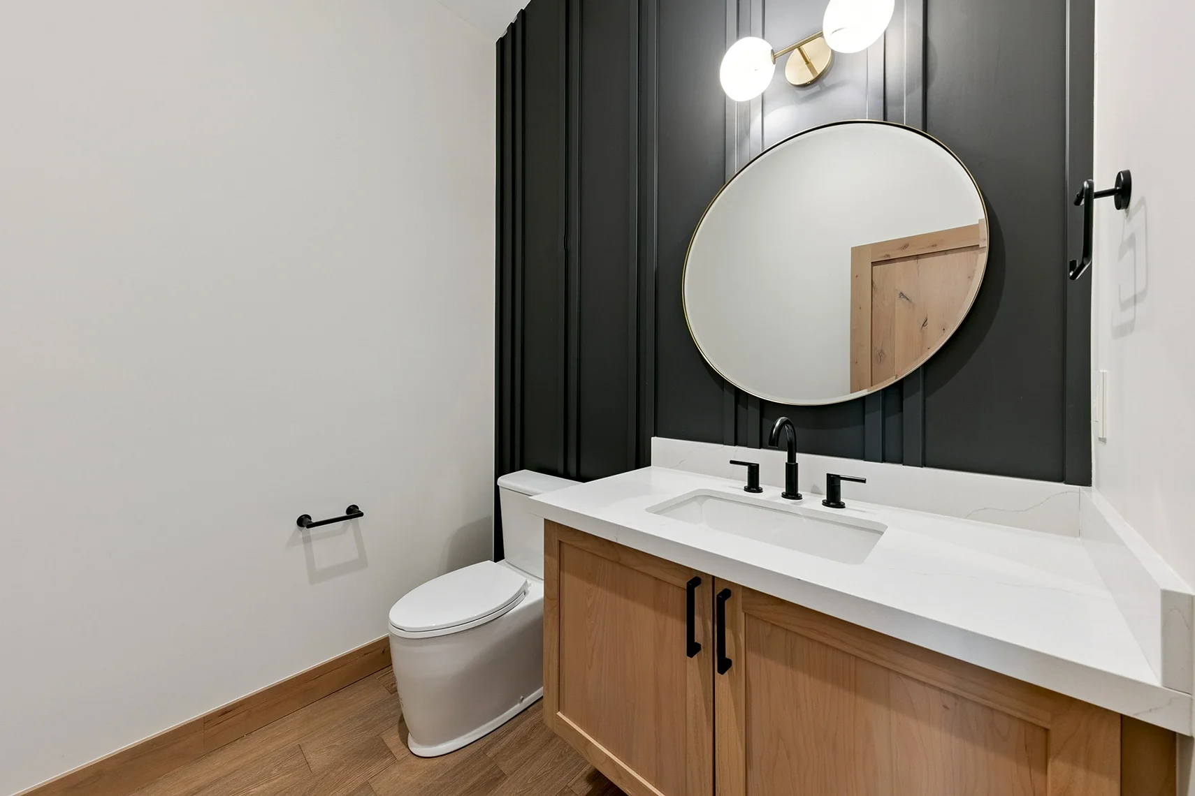 Modern bathroom with white walls, wood flooring, a black wood-paneled accent wall, and a round mirror.