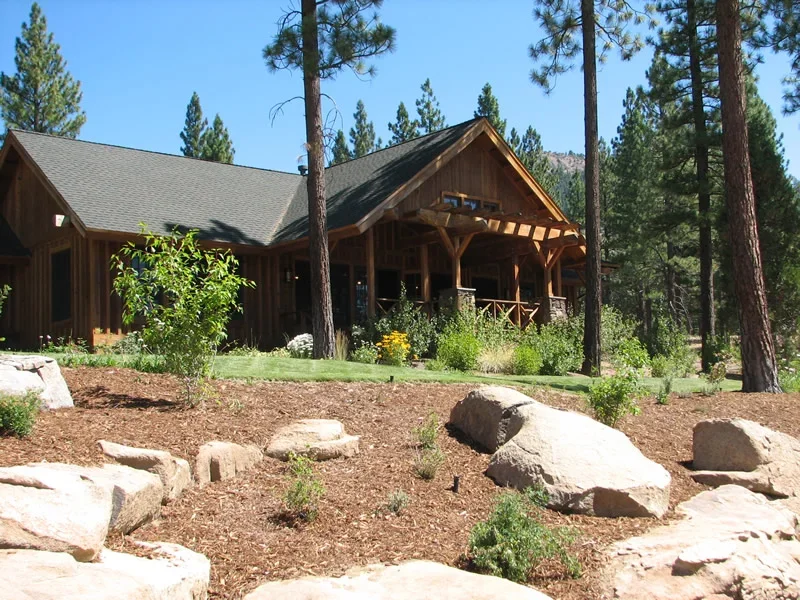 A wooden house with a pitched roof surrounded by tall pine trees and landscaped yard with rocks, bushes, and flowers.
