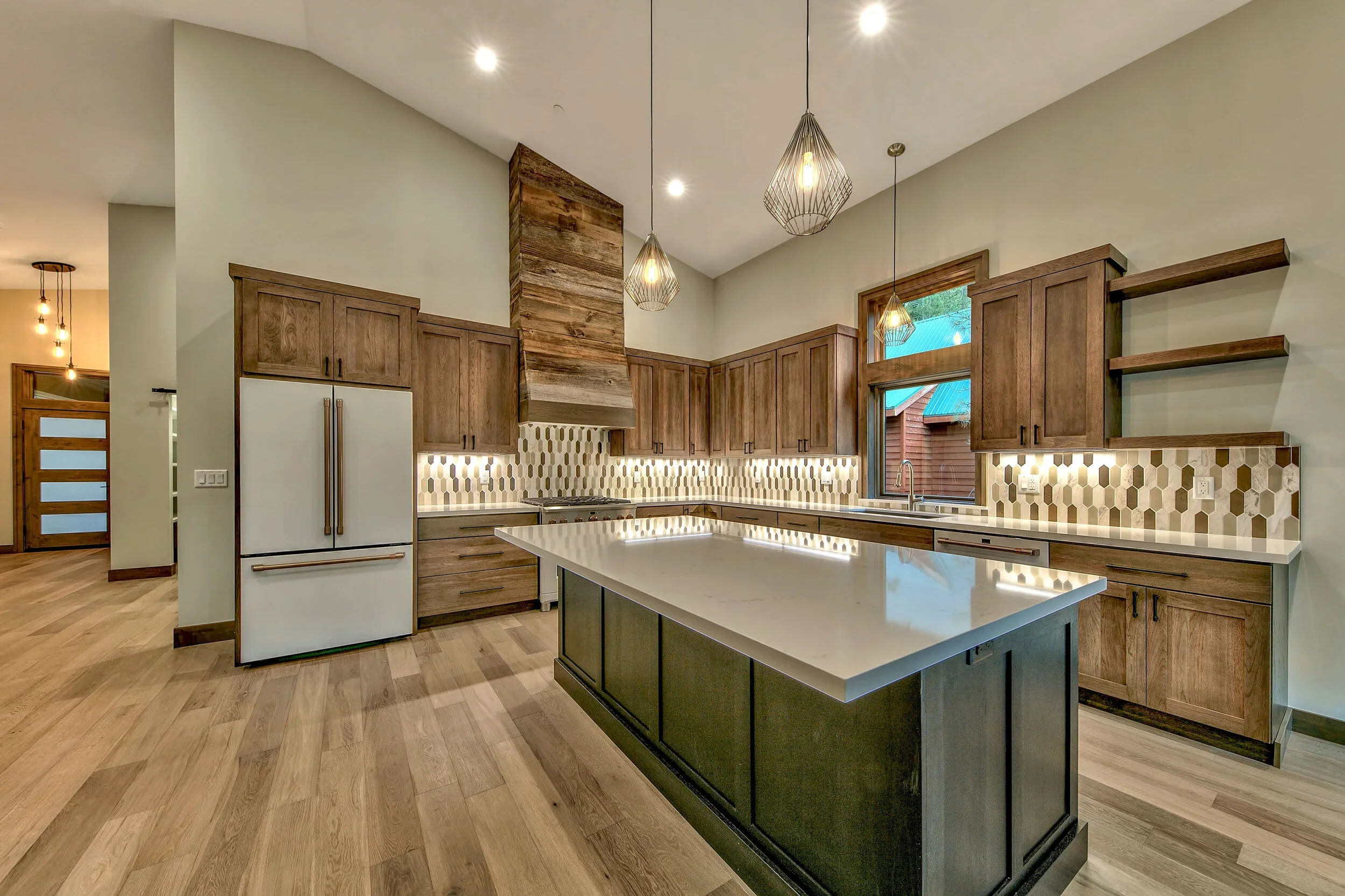 Modern kitchen with wooden cabinets, a white island with a dark base, geometric backsplash, large window, pendant lights, and open shelves.