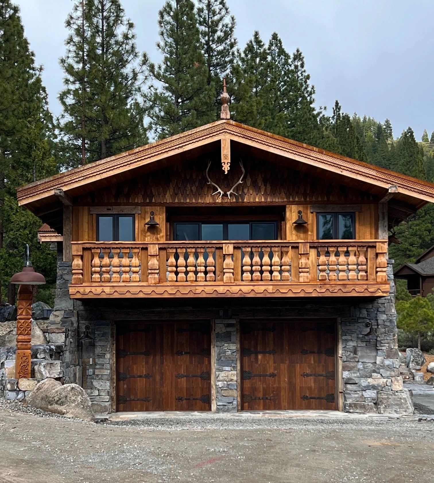 A two-story wooden house with stone accents, a balcony with carved wooden balustrades, and a pair of garage doors on the lower level. The house features decorative antlers above the balcony and a weather vane on the roof, surrounded by tall pine tree